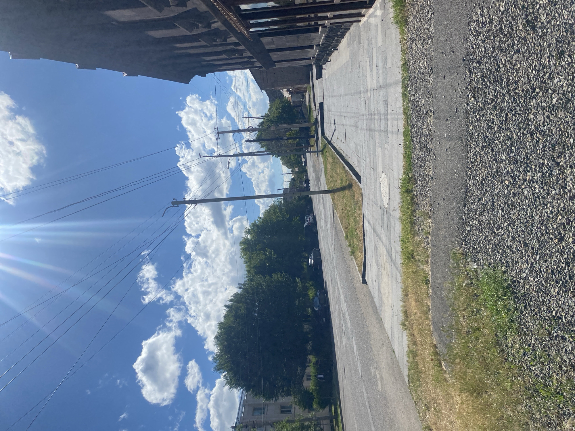 The image depicts an outdoor scene, likely a street or sidewalk area, under a bright, sunny sky with scattered clouds. The perspective is slightly tilted, suggesting the photo was taken from an unusual angle or the camera was not held level.
In the foreground, a paved pathway made of rectangular stones runs diagonally across the frame, bordered by a strip of dry grass and then a section of dark grey gravel. A road is visible to the left of the gravel area. Overhead, a complex network of electrical wires and poles dominates the upper left portion of the image, stretching across the blue sky. The sun is intensely bright, casting lens flare and streaks of light into the shot.
In the background, lush green trees line the side of the road, their dense foliage obscuring much of what lies behind them. Several dark-colored cars are parked along the roadside, partially visible between the trees. A building with a light-colored facade and visible windows can be seen at the far end of the street, peeking out from behind the trees. Above the street and trees, a large, dark structure with a repeating pattern of horizontal and vertical elements is visible, possibly an overpass or an elevated walkway.
The time of day appears to be daytime, likely late morning or early afternoon, given the strong sunlight. The weather is clear and sunny, indicated by the bright sky and the lens flare. There is no visible text in the image. The scene conveys a sense of an ordinary day in an urban or suburban environment.