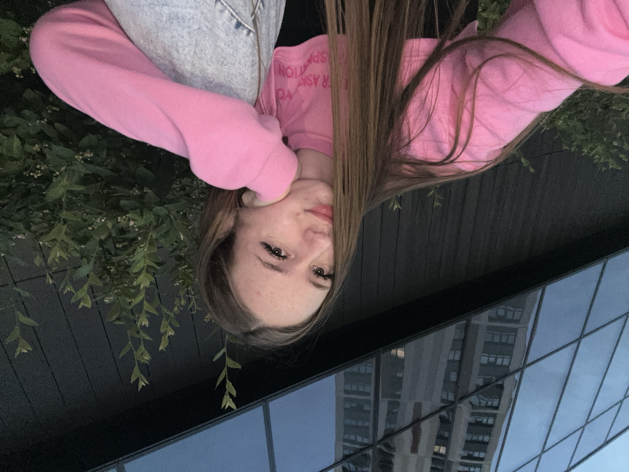 A young woman is pictured outdoors in a city setting, leaning her head on her hand. She has long, brown hair and is wearing a pink sweatshirt and jeans. Behind her, a modern building with a glass facade reflects the sky, and green bushes are visible. The scene appears to be taken at dusk or during twilight.