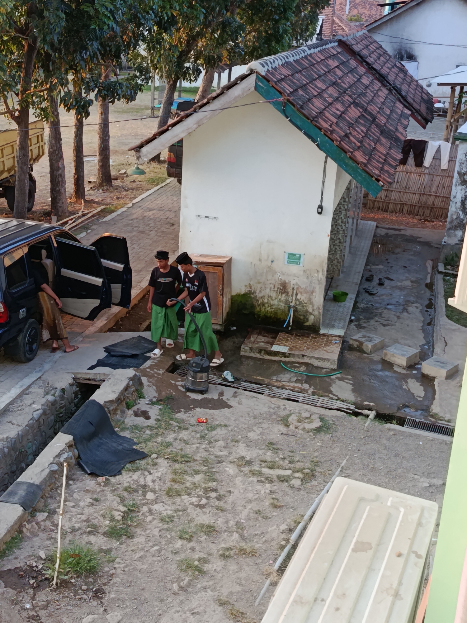 The image depicts an outdoor scene in Ramban Wetan, Indonesia, likely in a residential or communal area. Two young males, dressed in black t-shirts and green sarongs, are standing near a dark-colored SUV with its doors open. One of them is holding a vacuum cleaner hose, suggesting they might be cleaning the vehicle. Another person, partially visible inside the SUV, is wearing a tan sarong. In the background, a small building with a red tiled roof is present, and a truck is parked to the left. The ground is a mix of concrete, dirt, and some grass. A water tap and a green bucket are visible near the building, indicating a water source. The overall environment appears to be somewhat cluttered, with various objects scattered around.