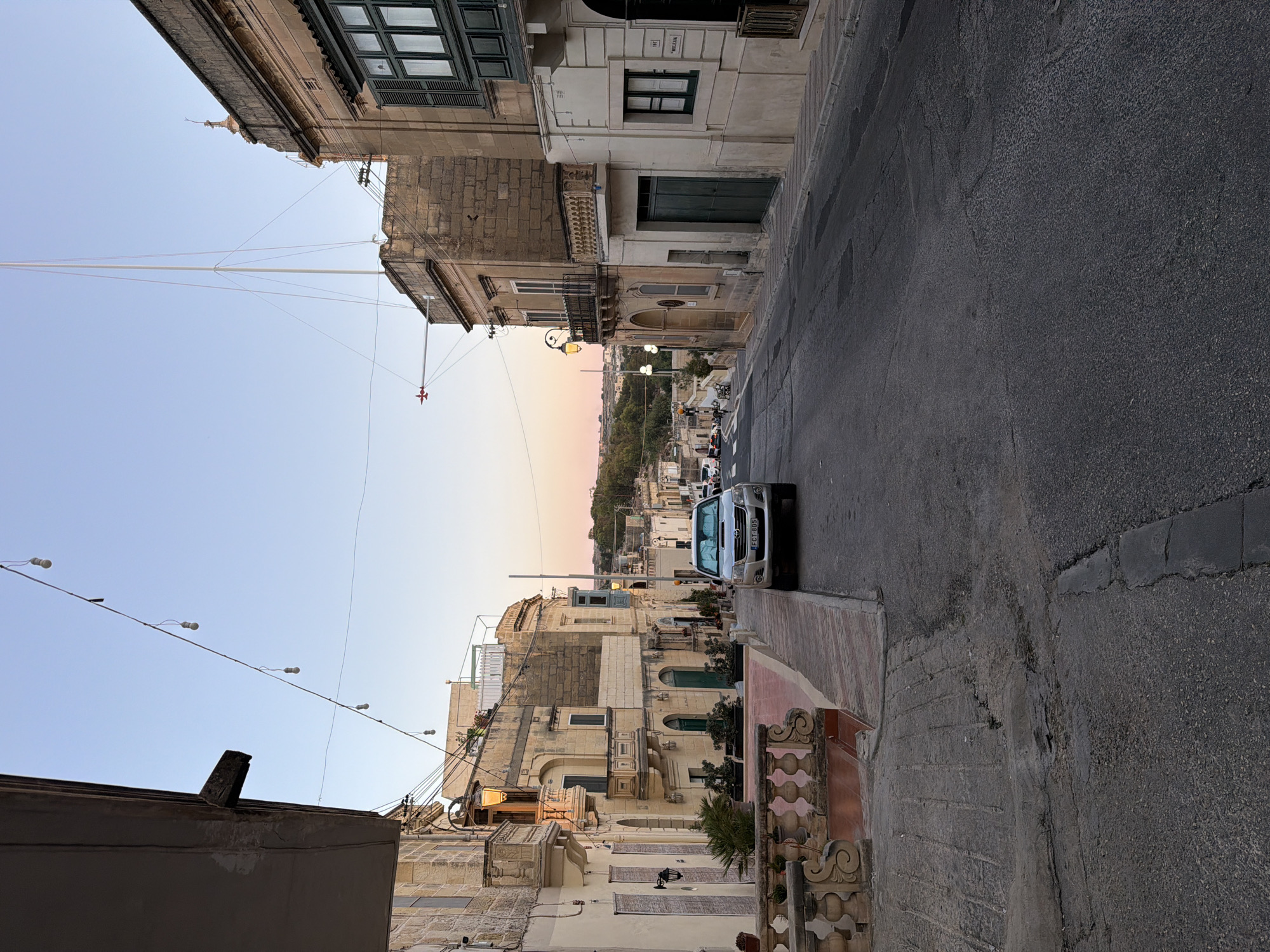 The photograph captures a street scene in Fontana, Malta, during what appears to be twilight. The perspective is from a slightly elevated position looking down a moderately sloped street. On the left, a building with a dark overhang juts into the frame, and below it, a stone balustrade with decorative elements leads to a paved area with planters. Further down the street on the left, traditional Maltese architecture is visible, characterized by stone buildings with balconies, some featuring ornate railings.
The street itself is asphalt, showing signs of wear and tear with visible cracks. A silver SUV is parked on the right side of the street, facing uphill, with its license plate clearly visible. The right side of the street is lined with buildings, also in typical Maltese style, featuring stone facades, balconies (one with dark green shutters), and arched doorways. String lights are suspended across the street above the buildings, with a few bulbs visible against the clear sky. A tall mast, possibly for a flagpole or antenna, rises from behind one of the buildings.
In the background, the street continues to curve uphill, with more buildings and hints of vegetation. The sky exhibits a soft gradient of blues and pinks, indicative of sunset or sunrise. The overall impression is a quiet, picturesque street in a Mediterranean town.