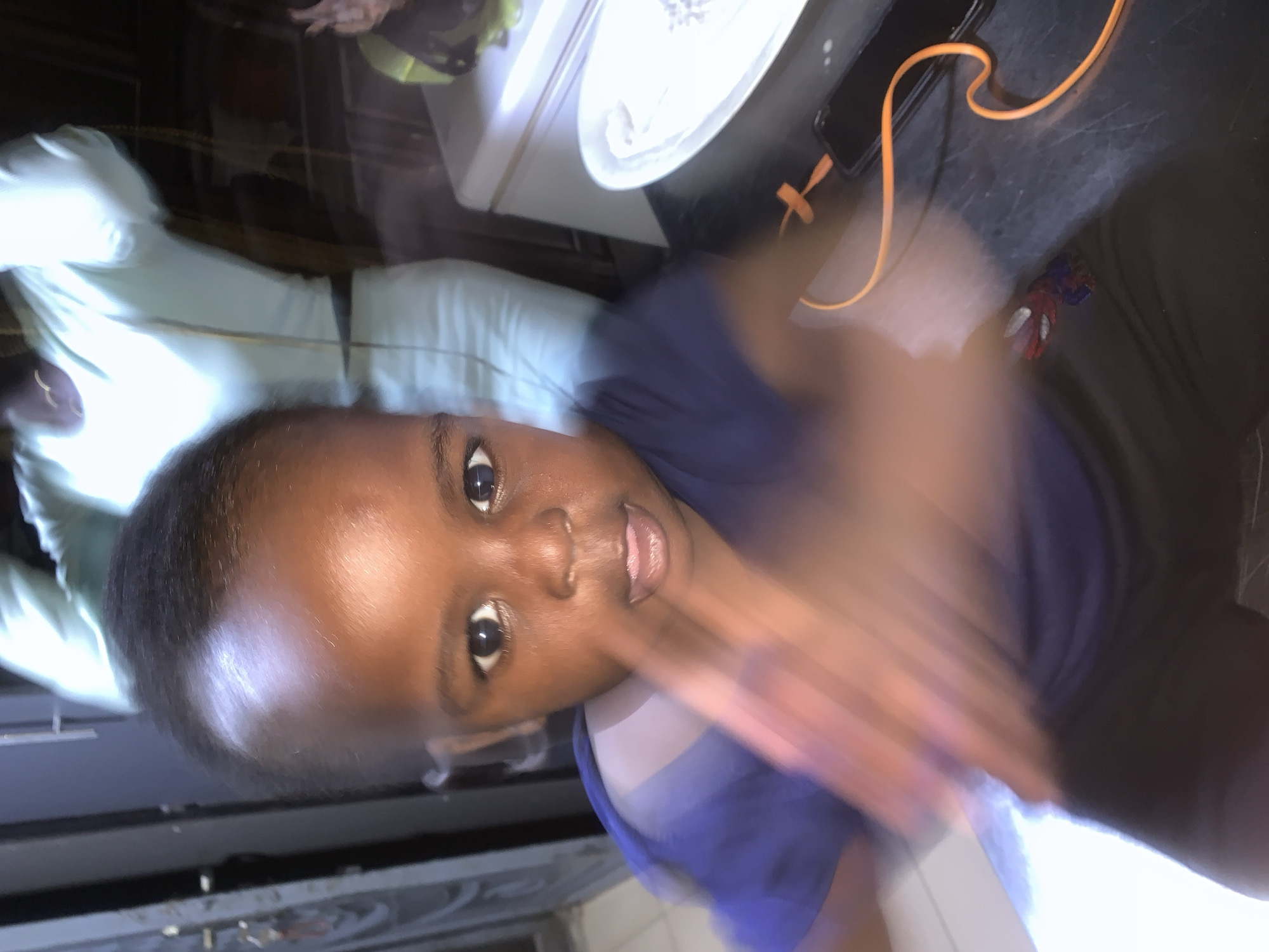 A young child, appearing to be around 1-2 years old, is the primary subject of this close-up, slightly blurry indoor photograph. The child has dark skin and black hair, with large, expressive eyes and a small mouth slightly ajar, revealing their tongue. They are wearing a dark blue t-shirt and dark pants. The child's right hand is blurred due to motion, positioned in front of their face as if interacting with the camera or an object.
In the background, a person, likely an adult, is visible, wearing a light blue garment and possibly a necklace. The environment appears to be a home, with what looks like a refrigerator or large appliance to the right of the frame, and a dark countertop surface where a smartphone and an orange charging cable are placed. There is also a white plate on the countertop. A door or cabinet is visible to the left of the frame. The lighting suggests it is indoors, potentially at night or in a dimly lit space. The overall impression is a candid, spontaneous moment captured in a domestic setting.