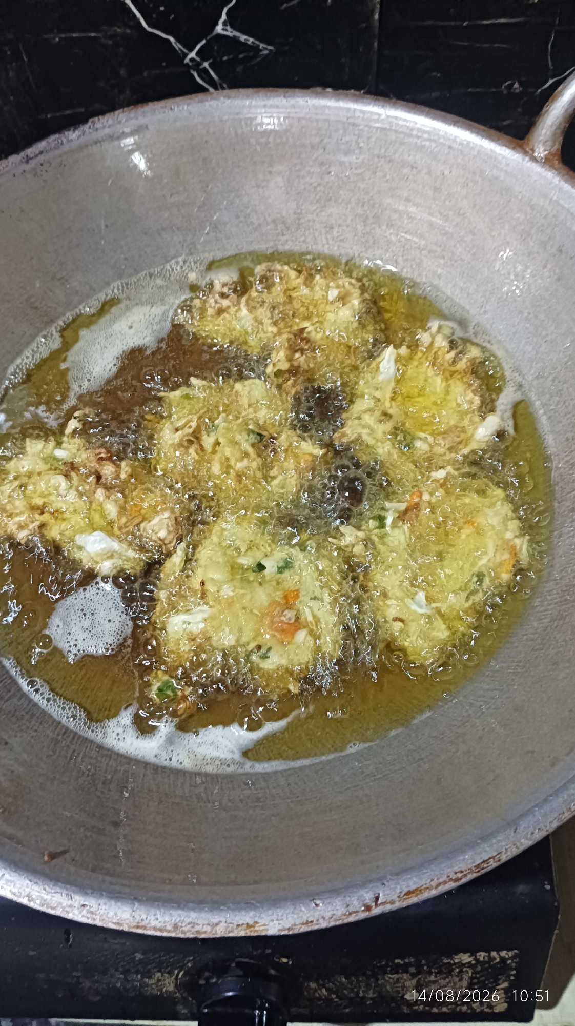 The image shows a close-up, overhead view of several fritters frying in a metal wok filled with hot oil. The fritters appear to be made from a mixture of vegetables and batter, and they are golden brown and bubbling as they cook. The wok is on a stovetop, and the background shows a black tiled wall. A timestamp in the lower right corner of the image reads "14/08/2026 10:51". There is no indication of the specific location in Tasikmalaya, Indonesia.