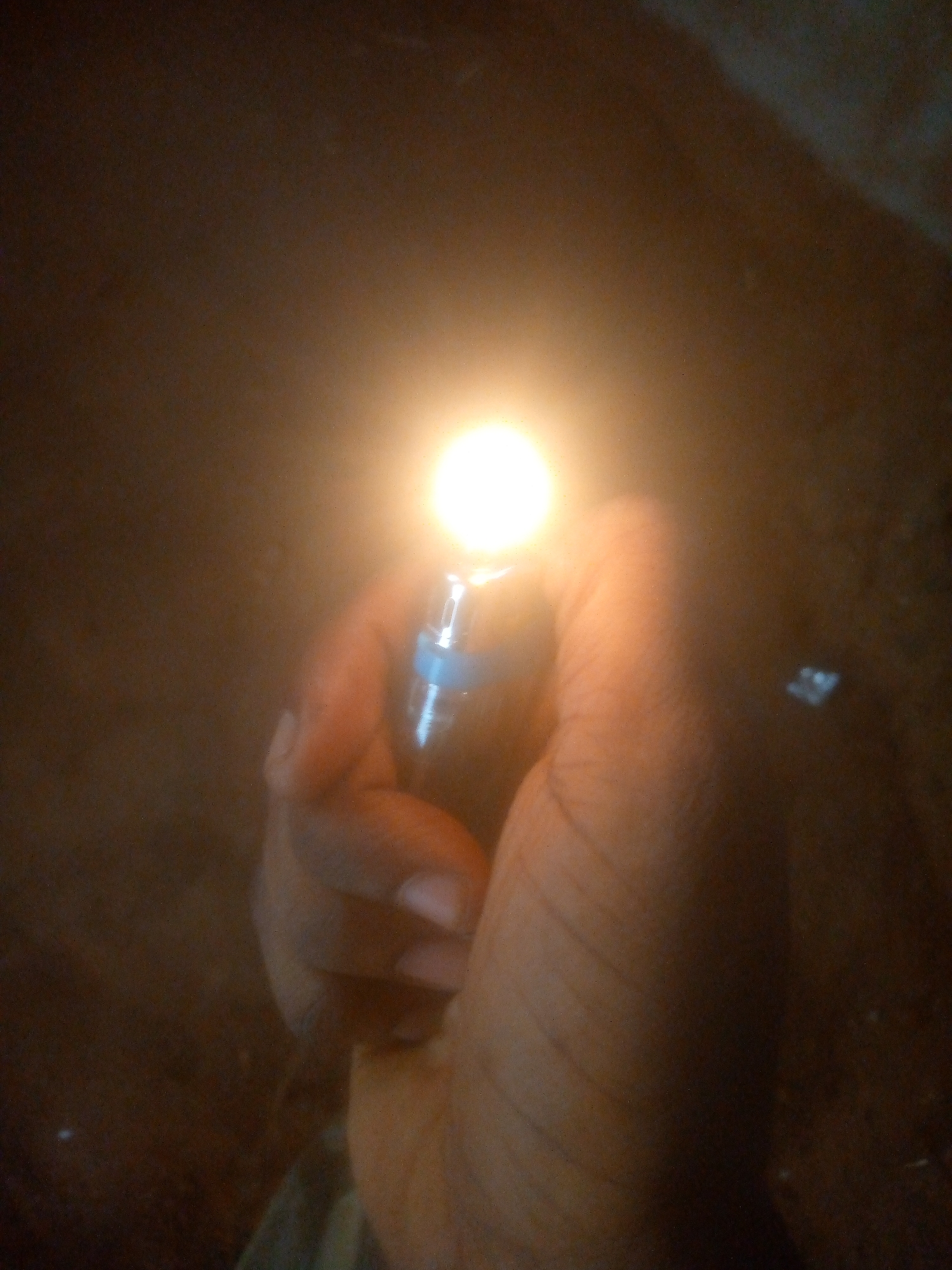 The image captures a close-up, first-person perspective of a human hand holding a lit lighter in a very dark environment. The bright, yellow-orange flame from the lighter is the dominant light source, illuminating the fingers and palm of the hand. The hand appears to be a right hand, with warm, brown skin tones. The lighter itself has a visible metallic or blue-tinted section just below the flame.
The background is almost entirely dark and out of focus, suggesting either nighttime or an extremely dim indoor setting. No specific details or objects are discernible in the background, only a general dark brown and black expanse, which occasionally catches subtle glints of light from the flame. The overall impression is one of a small, isolated source of light against an encompassing darkness. There is no visible text in the image.