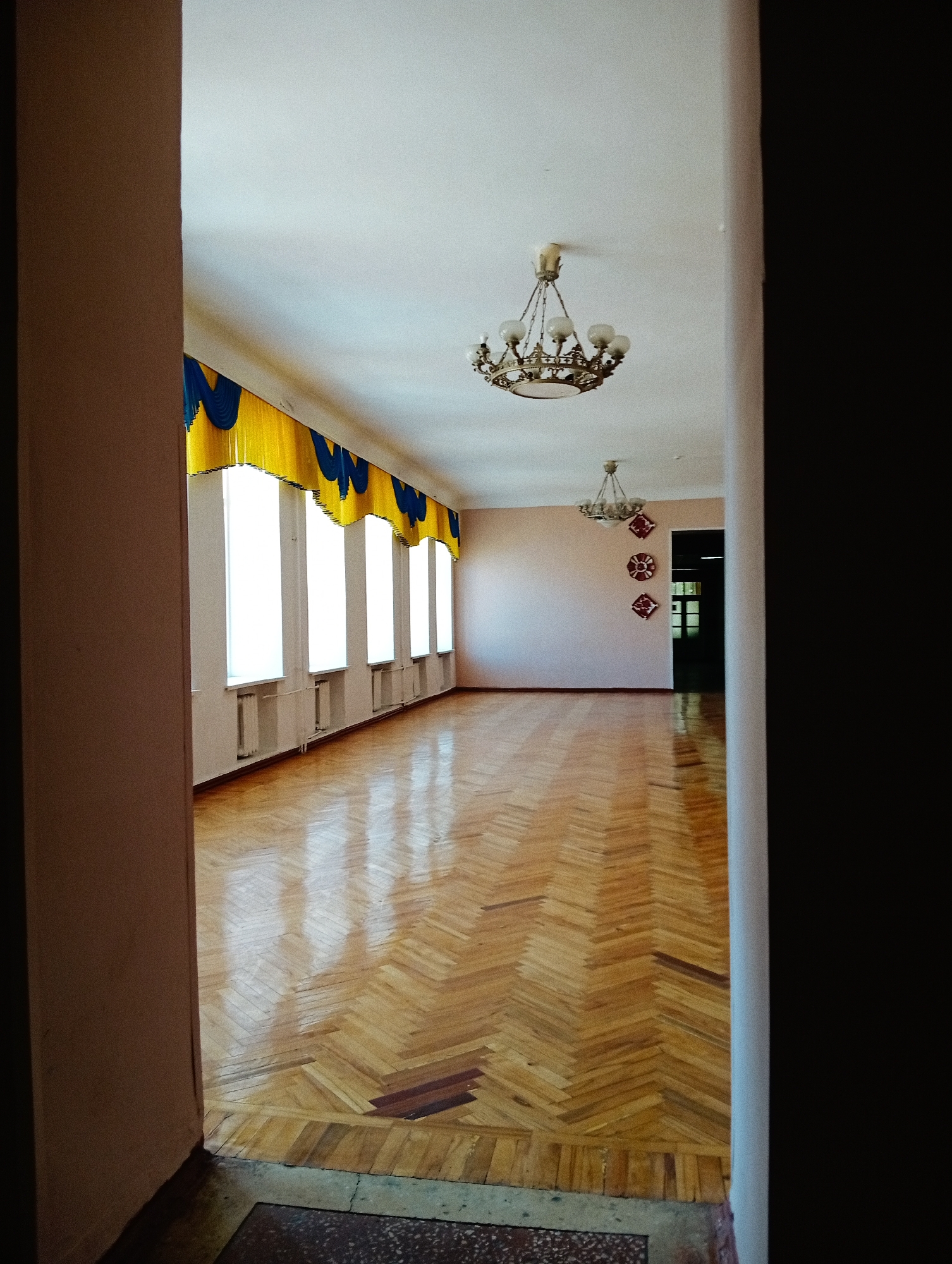 The image depicts a long, empty hall or room in Poltava, Ukraine, viewed from an entryway framed by two walls. The left wall of the entryway is a light brown, while the right wall is mostly in shadow, appearing very dark.
The room features a highly polished wooden parquet floor laid in a herringbone pattern, reflecting bright light streaming in from a series of five tall, narrow windows on the left side of the hall. Above these windows, decorative valances in bright yellow with blue scalloped details are hung, adding a touch of color. White radiators are visible beneath each window.
Two ornate chandeliers with multiple light bulbs hang from the white ceiling, casting soft light. The first chandelier is closer to the viewer, and a second, smaller one is positioned further down the hall. On the far wall, three decorative elements, possibly circular or square, are arranged vertically and feature intricate patterns, reminiscent of Ukrainian folk art. A doorway at the very end of the hall provides a glimpse into a darker interior space.
The walls are painted a light, neutral color, possibly a pale peach or beige. The room is entirely empty of people or furniture, suggesting a moment of quietude, perhaps before or after an event, or simply a vacant space. The bright illumination from the windows indicates it is daytime. There is no visible text in the image.