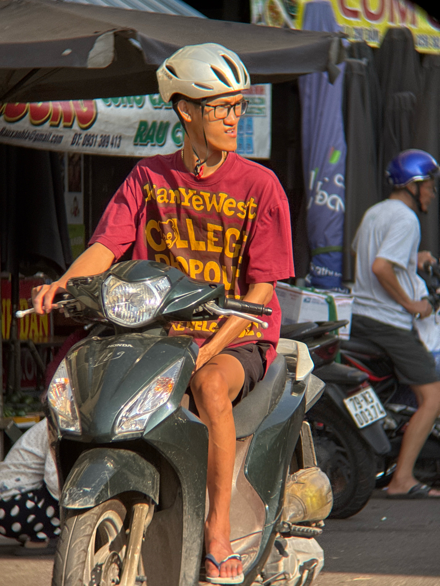 A young man wearing glasses and a red t-shirt with yellow text sits on a dark green scooter. He has a white helmet with black straps on his head. The scooter has a reflective chrome headlight. In the background, another person in a white shirt and blue helmet is visible, also on a scooter. Various stalls and awnings are set up along the street, typical of street vendors in Nha Trang, Vietnam. The scene appears to be a busy street market or shopping area.