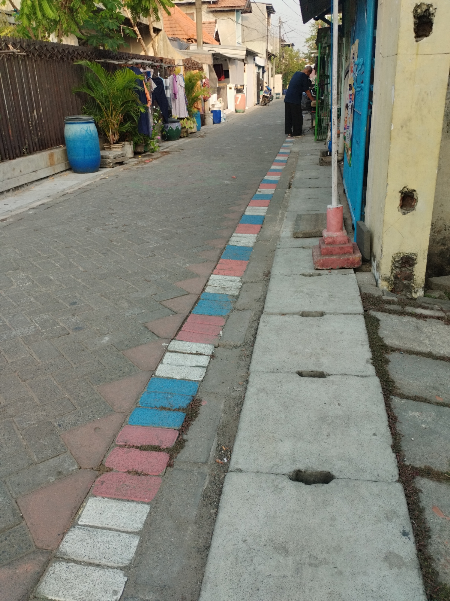 A narrow, paved alleyway runs through a residential area in Surabaya, Indonesia. The alley is lined with houses and fences. On the left side of the alley, a blue plastic barrel and several potted plants are visible. Clothes are hanging on a clothesline in front of a house. The alley is decorated with a border of red, white, and blue painted bricks. On the right side of the alley, a person in a blue shirt and black pants is working on something near a blue metal gate. A yellow-painted wall with damaged sections is next to the gate. Concrete slabs with openings form a drainage channel along the right side of the alley.