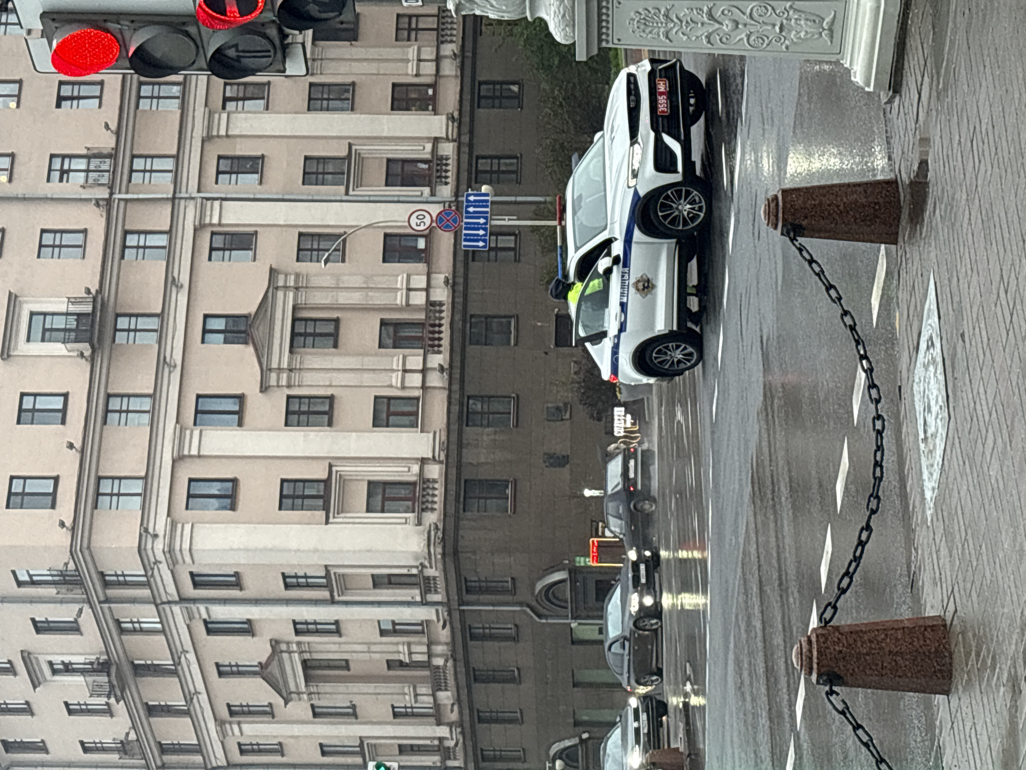 The image shows a street scene in Minsk, Belarus on a rainy day. A police car is parked on the right side of the street near a traffic light displaying a red signal. The car has license plate 3595 MH. A police officer is inside the car. Other cars are visible in the background along the wet road. A chain connecting stone bollards runs along the edge of the sidewalk. A large building with numerous windows is on the left side of the street. A speed limit sign (50) is visible next to the police car. The scene is urban, with a focus on traffic and architectural details.