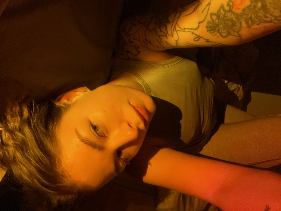 A young woman reclines, illuminated by warm light, her face angled towards the camera. She wears a light green tank top. Her hair is pulled back. Tattoos adorn both arms. The background is muted and dark.