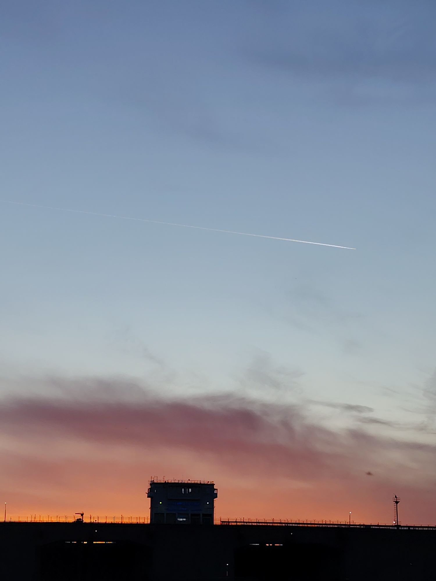 The image depicts a structure, possibly a building or control tower, situated on what appears to be a bridge or dam. The structure is dark in silhouette against a backdrop of a sunset sky. The sky transitions from orange at the bottom to blue at the top, with a pinkish-purple cloud formation near the horizon. A contrail from an airplane is visible in the upper portion of the sky. The location is in Chaikovsky, Russia.
