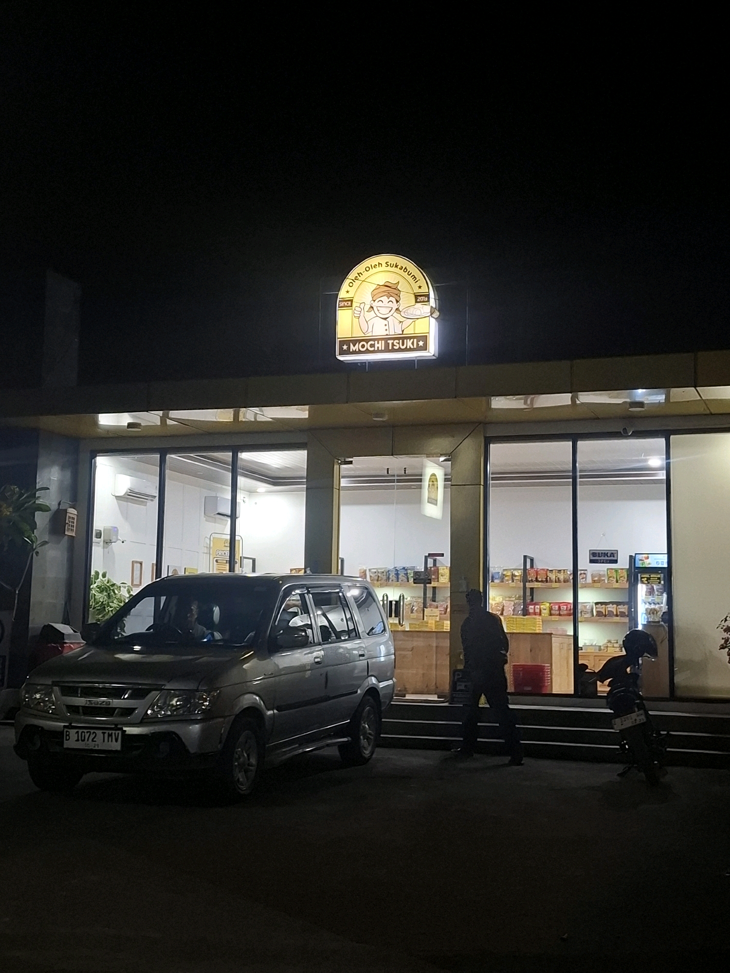 In Karangtengah, Indonesia, at night, a silver SUV is parked in front of a brightly lit storefront. Above the entrance, a yellow neon sign reads "MOCHI TSUKI" and features a cartoon character. The storefront windows reveal shelves stocked with products. A man stands outside the store, silhouetted against the interior light, and a motorcycle is parked to the right of the entrance. The license plate on the SUV reads "B 1072 TMV".