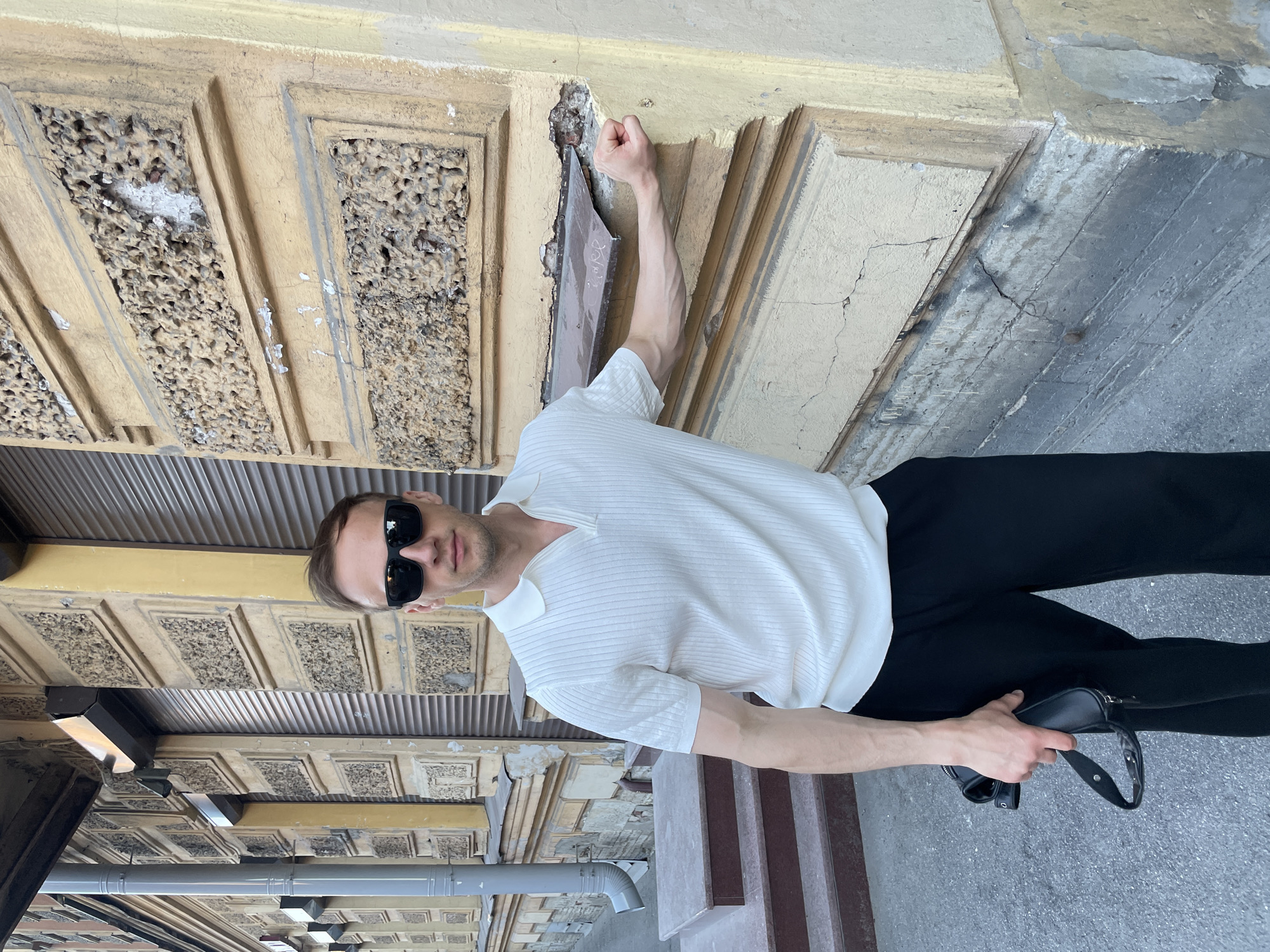 An adult male stands on a sidewalk in Saint Petersburg, Russia, in front of an aged building facade. The man, with short brown hair, wears dark rectangular sunglasses, a white ribbed short-sleeved polo shirt with a V-neck zipper, and black trousers. He is positioned with his right elbow and fisted hand leaning against a yellow/ochre painted building column, revealing prominent veins on his forearm. In his left hand, he holds a small black zippered bag with a strap.
The building behind him exhibits typical Saint Petersburg architecture, featuring a faded yellow/ochre painted plaster exterior with decorative rectangular panels. These panels are constructed with a rough, textured stucco that shows significant deterioration, including chipping, peeling paint, and exposed underlying material. Dark grey roll-down shutters cover openings on the building's ground level. A grey metal downspout runs vertically on the left side of the facade, adjacent to a set of three brown steps. The lower section of the building's corner displays exposed grey material with cracks and signs of wear. The ground is an asphalt surface. The scene is illuminated by diffused daylight.