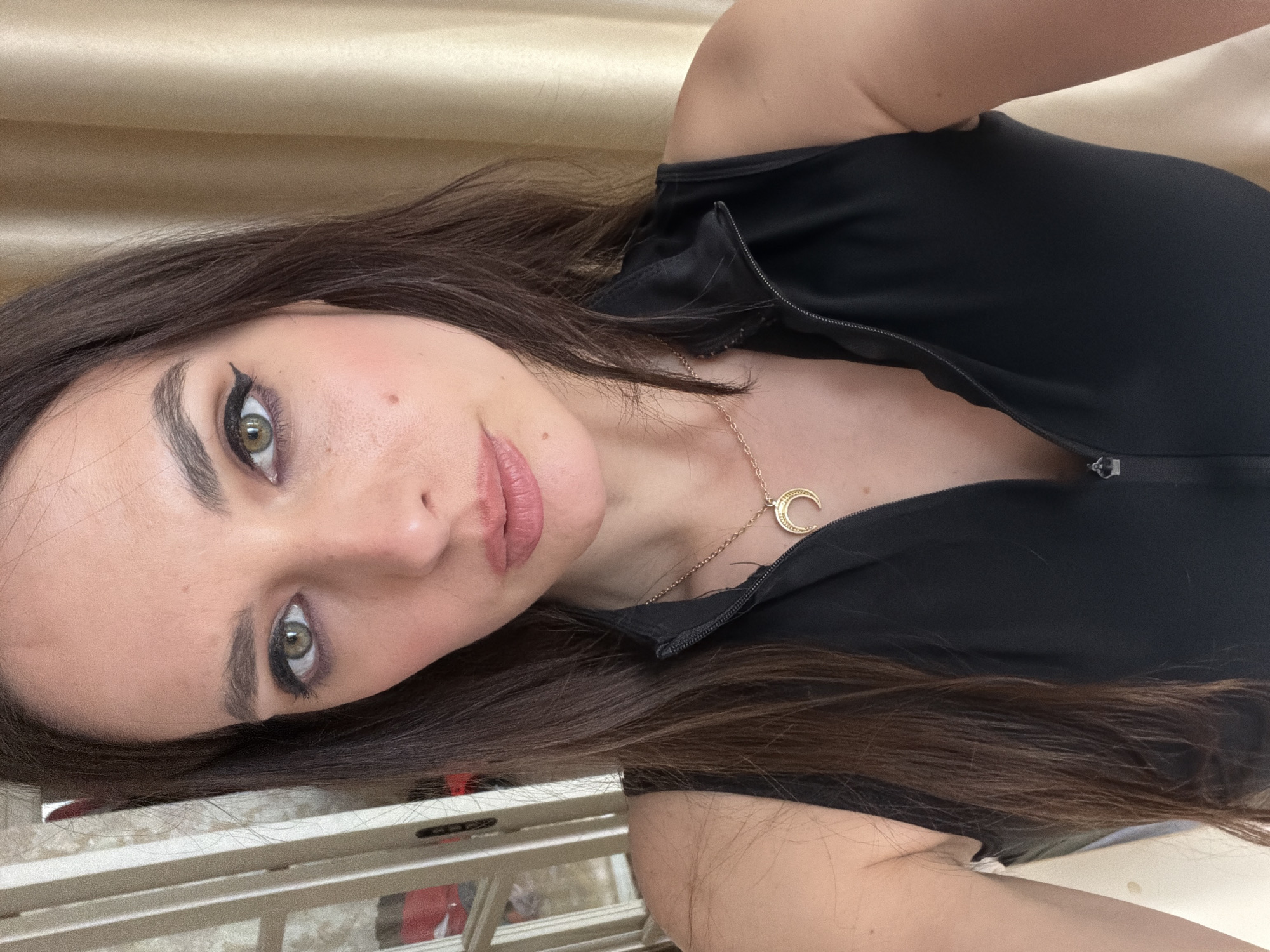 The media file is a selfie of a young woman in Ufa, Russia. She has long, dark brown hair and light green eyes. She is wearing a black top with a zipper and a gold necklace with a crescent moon pendant. The background appears to be a room with a beige, textured wall and a window with a view of what looks like a street scene. The lighting suggests it is daytime. She has makeup on, including winged eyeliner and pink lipstick. Her expression is neutral. No text is visible in the image.