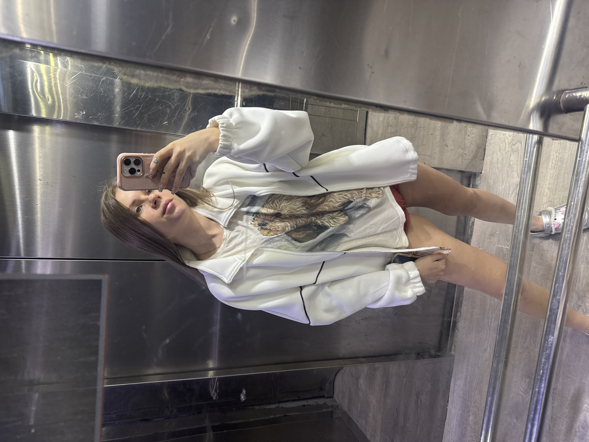 A woman is taking a selfie in what appears to be an elevator. She is wearing a white jacket over a t-shirt with a dog graphic. Her shorts are red and her footwear are silver slides. The elevator has stainless steel walls and a metallic railing. The photo may have been taken in Batumi, Georgia but that cannot be confirmed.