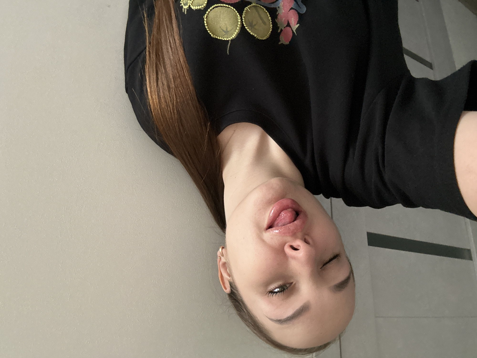 A woman with light skin and long, straight, brown hair is posing for a selfie. She is wearing a black long-sleeved shirt with a floral design in gold, pink, and blue. She is sticking her tongue out and winking. The background is a light gray wall with what appears to be a door or other architectural element visible in the upper right corner. The location is Chaikovsky, Russia.