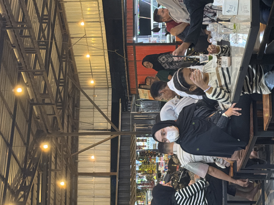 The image depicts an evening scene within an indoor or semi-outdoor dining establishment, likely a cafe or food court, located in Beurandang, Indonesia. The venue features an exposed structural ceiling composed of metal trusses supporting a corrugated metal roof, from which multiple bare incandescent light bulbs are suspended, providing illumination.
In the foreground, a young female child, wearing a striped long-sleeve shirt, jeans, and a silver headband, is seated at a dark-colored rectangular table. She holds a clear plastic cup containing a light-colored beverage to her mouth with both hands, actively drinking. Beside her cup on the table is another clear plastic cup holding a green liquid with a black straw, a clear plastic water bottle, and a white tissue dispenser branded "LIVI." To the child's left, a woman is seated on a wooden bench, wearing a black hijab, black clothing, and a white disposable face mask, looking slightly towards the right.
The midground and background are populated by numerous individuals, adults and possibly children, seated at similar tables and benches or standing. Visible individuals include a person in a black-and-white striped shirt and black hijab on the far left, partially obscured. A man in a white collared shirt and another man wearing glasses and a dark green shirt are visible in the mid-background. A woman in a patterned hijab is also observed further back, looking towards the right.
The environment includes dark wooden benches and dark-topped tables. In the far left background, a retail display or shelving unit holds various packaged goods. Red-painted horizontal metal beams or panels are visible in the mid-background, along with a partial sign displaying "BSI." The overall scene portrays a bustling social or dining gathering.