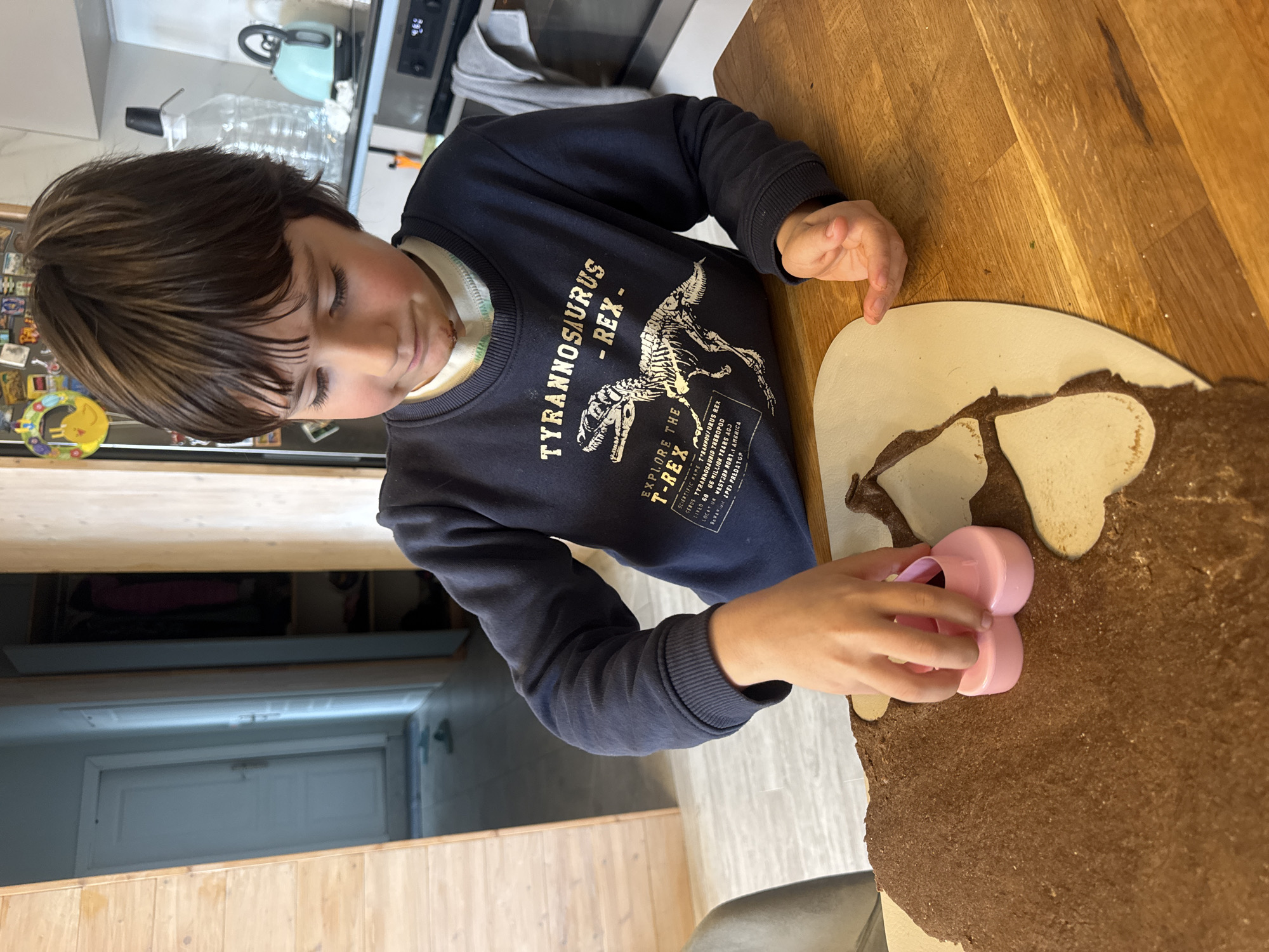 A young boy is making cookies on a wooden table. He is using a pink cookie cutter, shaped like a heart, to cut out cookies from a brown dough. He is wearing a navy blue sweatshirt with a dinosaur design on it. The environment appears to be a kitchen, with a refrigerator and other kitchen appliances visible in the background. There is no information about Burgas, Bulgaria.