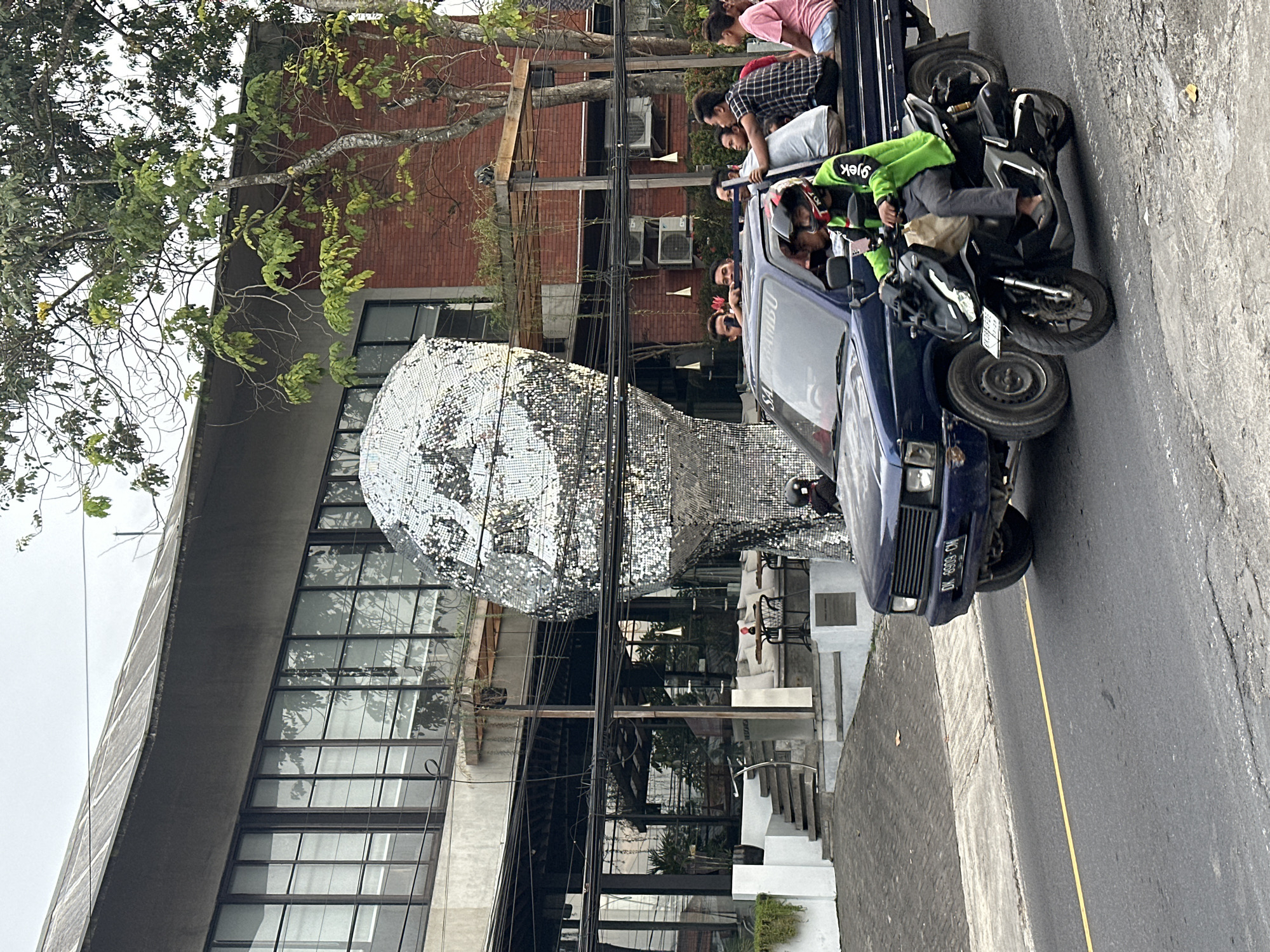 The photo depicts a scene in Ungasan, Indonesia. A blue car on the right side of the street is crowded with people, some leaning out of the car. A motorbike is loaded on top of the car's hood. On the left side of the road is a building with large windows and a mirrored, sculpture-like structure in front of it. Power lines run across the front of the building. The road has a yellow line marking the lanes.
