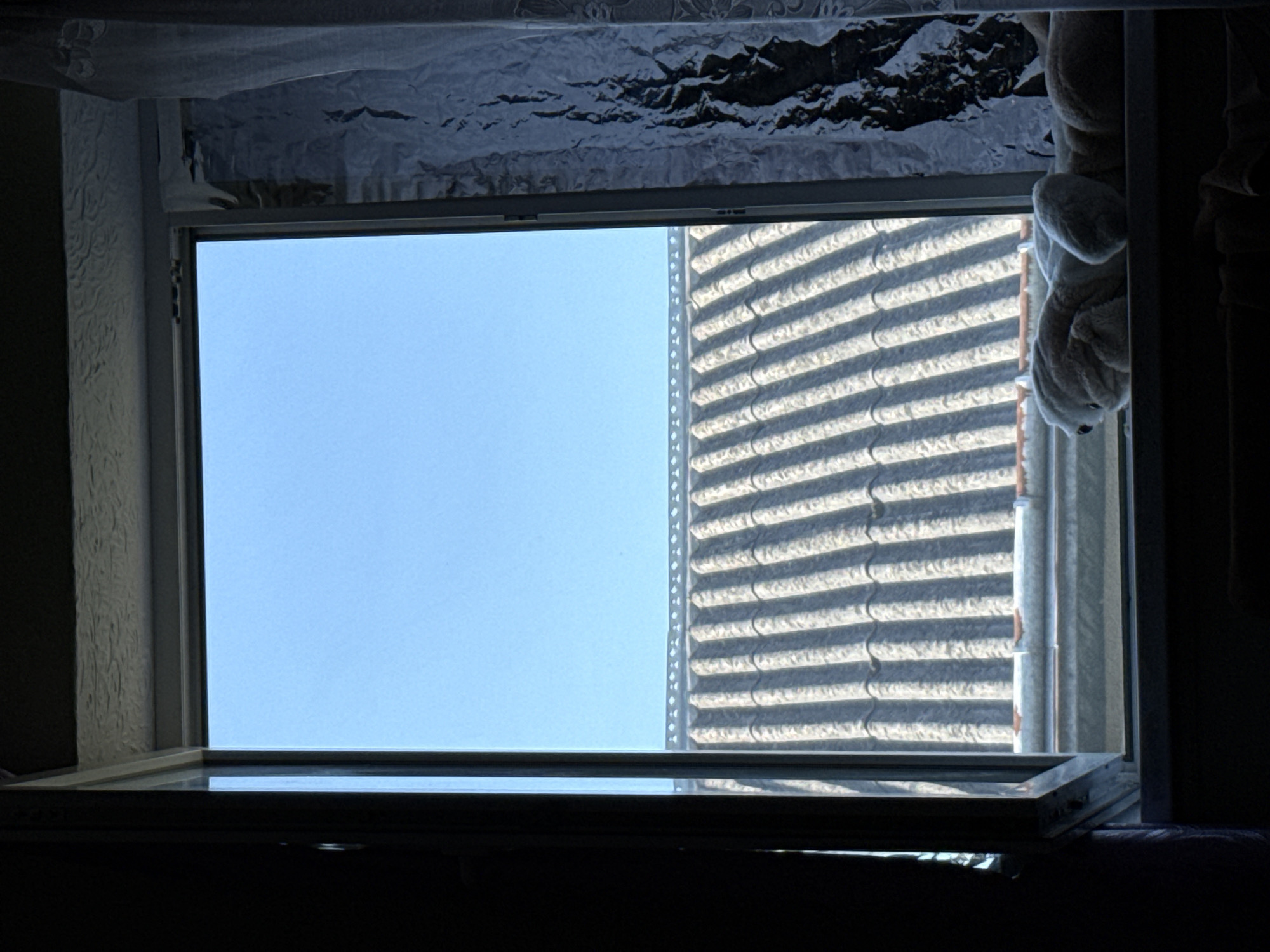 An open window reveals a clear blue sky above a row of grey, corrugated roof tiles. A white plush toy rests on the windowsill. The scene is framed by dark shadows to the left and a sheer white curtain to the right, which is textured and patterned. The bottom of the image shows a dark interior space. The image appears to be taken from inside a room.