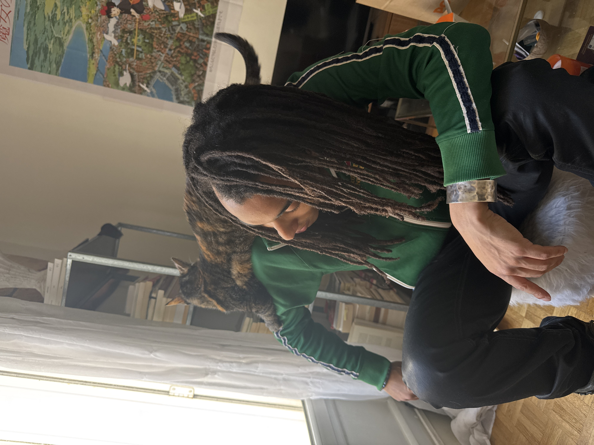 A person with dreadlocks is crouched near a window. The person wears a green jacket with white and navy stripes on the sleeves and black pants. A tortoiseshell cat sits on the person's back. A silver cuff is visible on the person's wrist. The window has white curtains. Behind the person is a metal shelf filled with books. A poster depicting a cityscape hangs on the wall, possibly a depiction of Paris, France, given the typical architecture. The floor appears to be made of wooden planks.