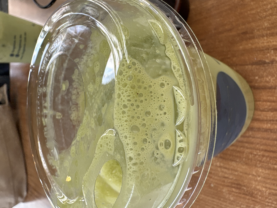 An overhead shot reveals a partially consumed green smoothie in a clear plastic cup with a dome lid. The smoothie is thick, with a frothy, bubbly texture visible through the lid, especially around the edges and within the circular opening. A blue or gray band is on the side of the cup. The container sits on a wooden surface, with a blurred light green object in the background.