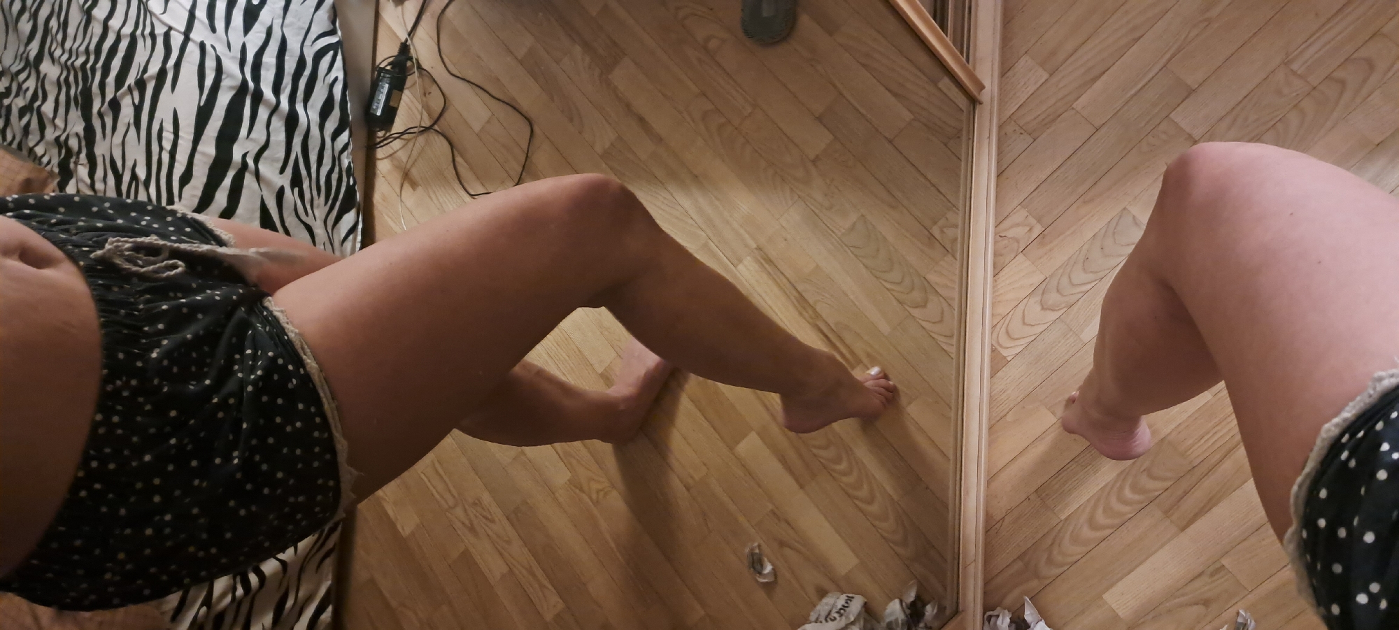 The image shows a first-person view of a person's body and legs. The person is wearing black shorts with white polka dots. The lower part of the body is visible along with the legs extended on a parquet floor. Next to the legs is a mirror. The reflection of the leg is visible in the mirror. A zebra-striped bedspread is partly visible in the upper-left part of the frame. In the background, there is an outlet with a power adapter. The lighting is bright. There is no clear evidence to suggest this is in Minsk, Belarus.