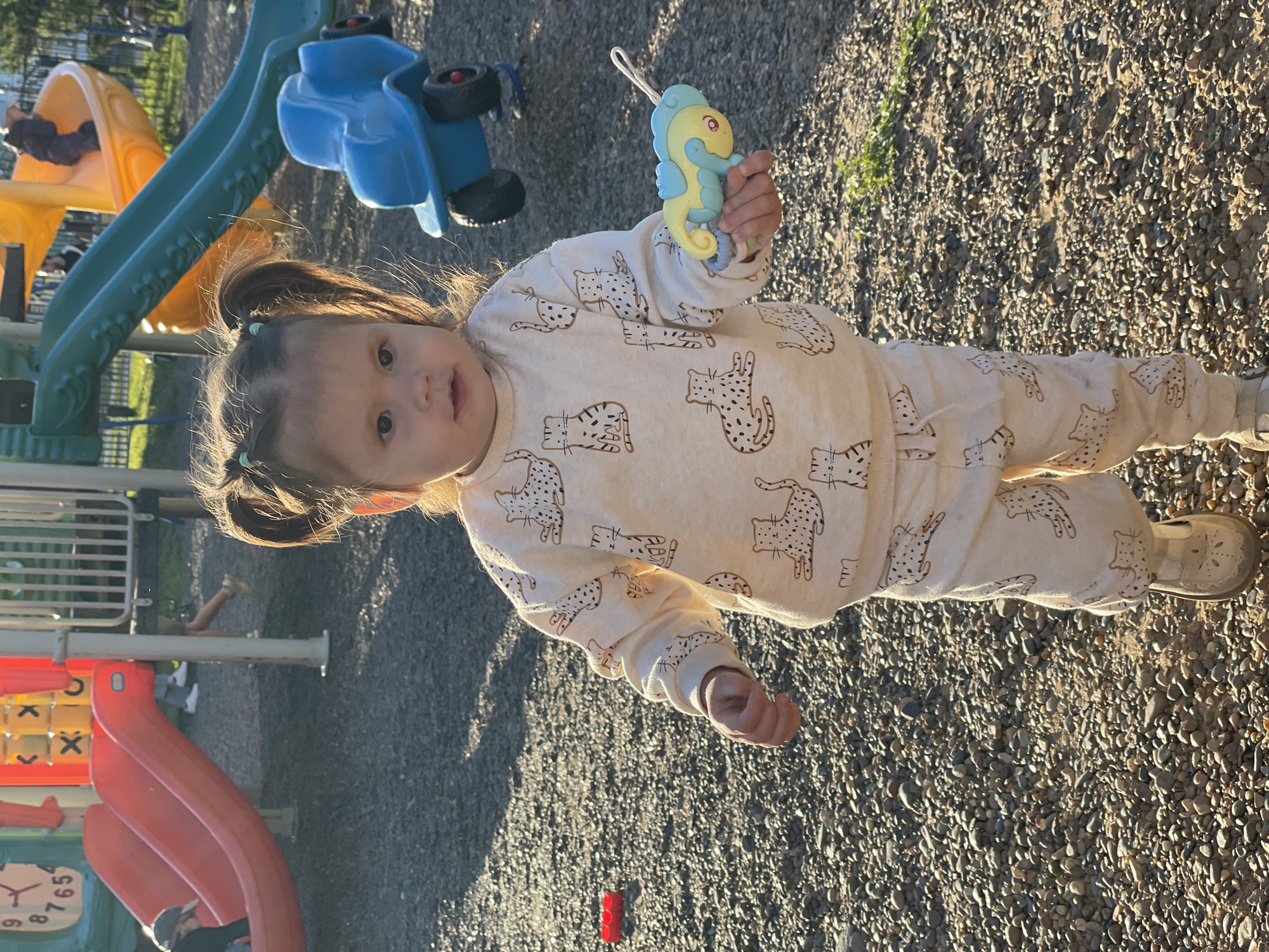 A toddler stands on a playground with a gravel surface. The child wears a light-colored pajama set patterned with tiger illustrations and holds a toy seahorse with a blue mane and yellow body in their right hand. In the background, playground equipment includes a green and yellow slide and a blue toy vehicle mounted on springs. A red plastic toy is visible on the ground near the toddler's feet. Other playground structures, including a red slide with a clock face, are also present in the background. The setting appears to be outdoors in daylight with dappled sunlight and shadows. There is no indication that the location is in Izhevsk, Russia.