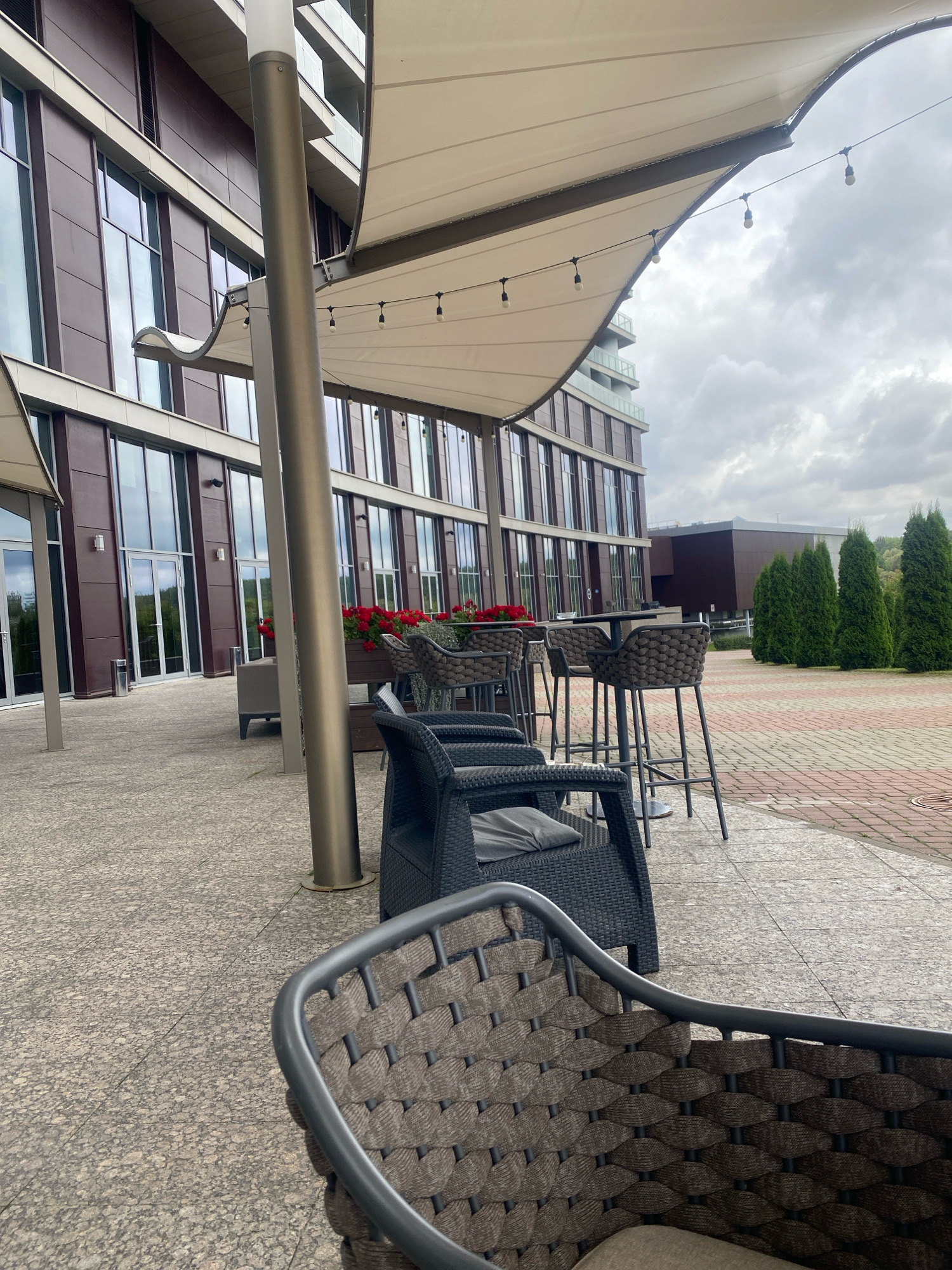 An outdoor seating area features woven furniture, including chairs and bar stools, arranged around dark tables. A large, beige awning, supported by a metal pole, provides shade. Behind the seating area, a modern building with dark brown paneling and numerous windows stands. The ground is paved with speckled gray tiles and a red and gray brick pattern. In the background, tall, conical evergreen trees line the property. The sky is overcast. The image appears to be taken in the city of Minsk, Belarus.