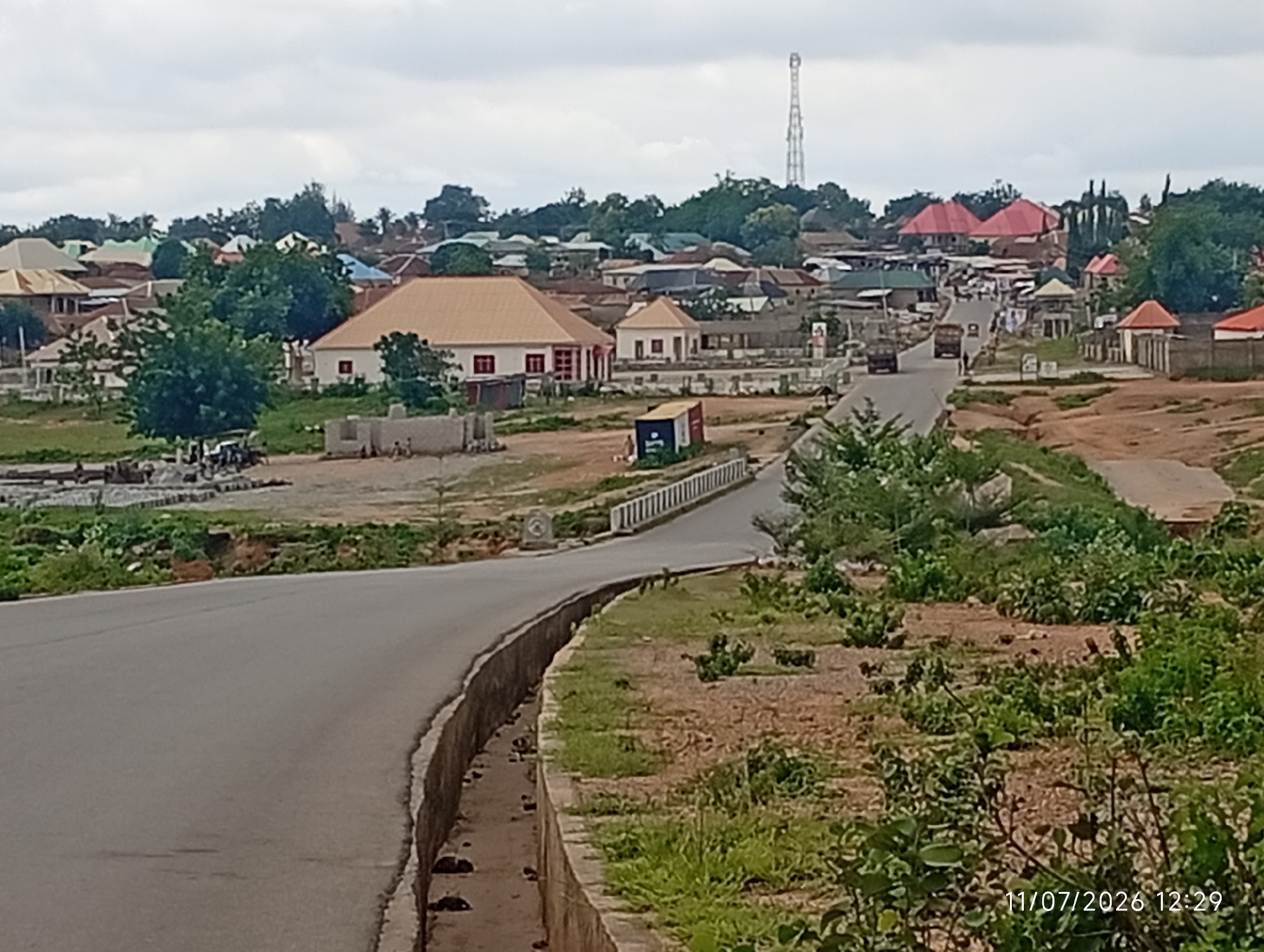 A scene in Uke, Nigeria, depicts a road curving through a town with buildings of various styles and colors. The sky is overcast with a pale, diffused light. In the foreground, a gray asphalt road curves to the left, bordered by a concrete retaining wall with an open drainage channel. Vegetation, including grass and shrubs, grows along the embankment.
Further back, across the road, are several structures. A large building with a prominent tan roof stands near the center, flanked by smaller buildings with red and blue roofs. A tall communication tower is visible in the distance, on a hill behind the town. Several vehicles, including trucks and a smaller car, are present on the road and in parking areas. People are visible in small groups, some near buildings and others on the road. Construction materials, possibly blocks and rebar, are piled to the left of the road. The overall impression is of a developing or established urban area within a rural or semi-rural setting. A timestamp "11/07/2026 12:29" is present in the lower right corner.