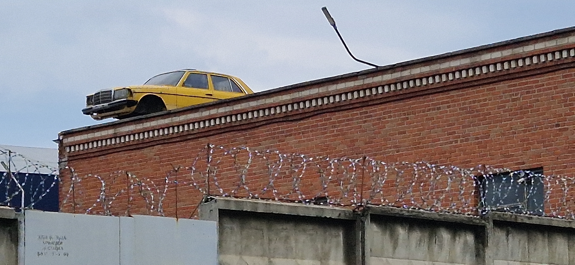 A yellow sedan is parked on top of a brick building, with its front wheels hanging over the edge. The car appears to be a vintage Mercedes-Benz. The building is topped with a decorative white border and a light fixture is positioned on the roof. Barbed wire is stretched along the top of a concrete wall in the foreground. The sky is overcast and a blue and white striped structure is visible to the left of the building. The scene is likely an urban industrial area.