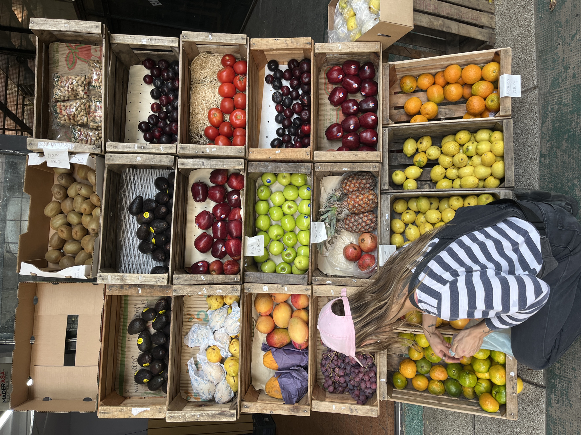 An overhead shot captures a vibrant outdoor produce stall, likely in Buenos Aires, Argentina, under bright daylight. A wide array of fresh fruits and vegetables is neatly displayed in wooden crates arranged on the ground.
The crates contain a diverse selection: peanuts packaged in clear bags, dark red plums, bright red tomatoes, and oranges are visible in the upper sections. Below them are green kiwis, dark avocados, and shiny red apples, some still bearing brand stickers. Other crates showcase vibrant green apples, whole pineapples, and reddish-orange pomegranates. Towards the bottom, yellow quinces or pears are individually wrapped in white paper, alongside ripe mangoes, papayas wrapped in purple paper, and bunches of dark red grapes. The crate closest to the visible person holds a mix of green and orange citrus fruits, possibly limes or a type of orange.
A person with long, light brown hair, dressed in a white and dark-striped long-sleeve shirt, black pants, and a pink baseball cap, is leaning down over one of the lower crates. Their hands are engaged with the citrus fruits, suggesting they are selecting items for purchase.
Visible text on small white signs indicates product names and prices: "Mani 12.000" (peanuts), "Kiwi 12.000 kg.", and "Naranja 9500 kg." (oranges). Some avocado stickers show "Agroideas," and box lining reads "ARGENTINAS." An empty cardboard box on the left is labeled "MADERASA." The scene depicts a bustling market activity, highlighting the abundance of fresh produce.