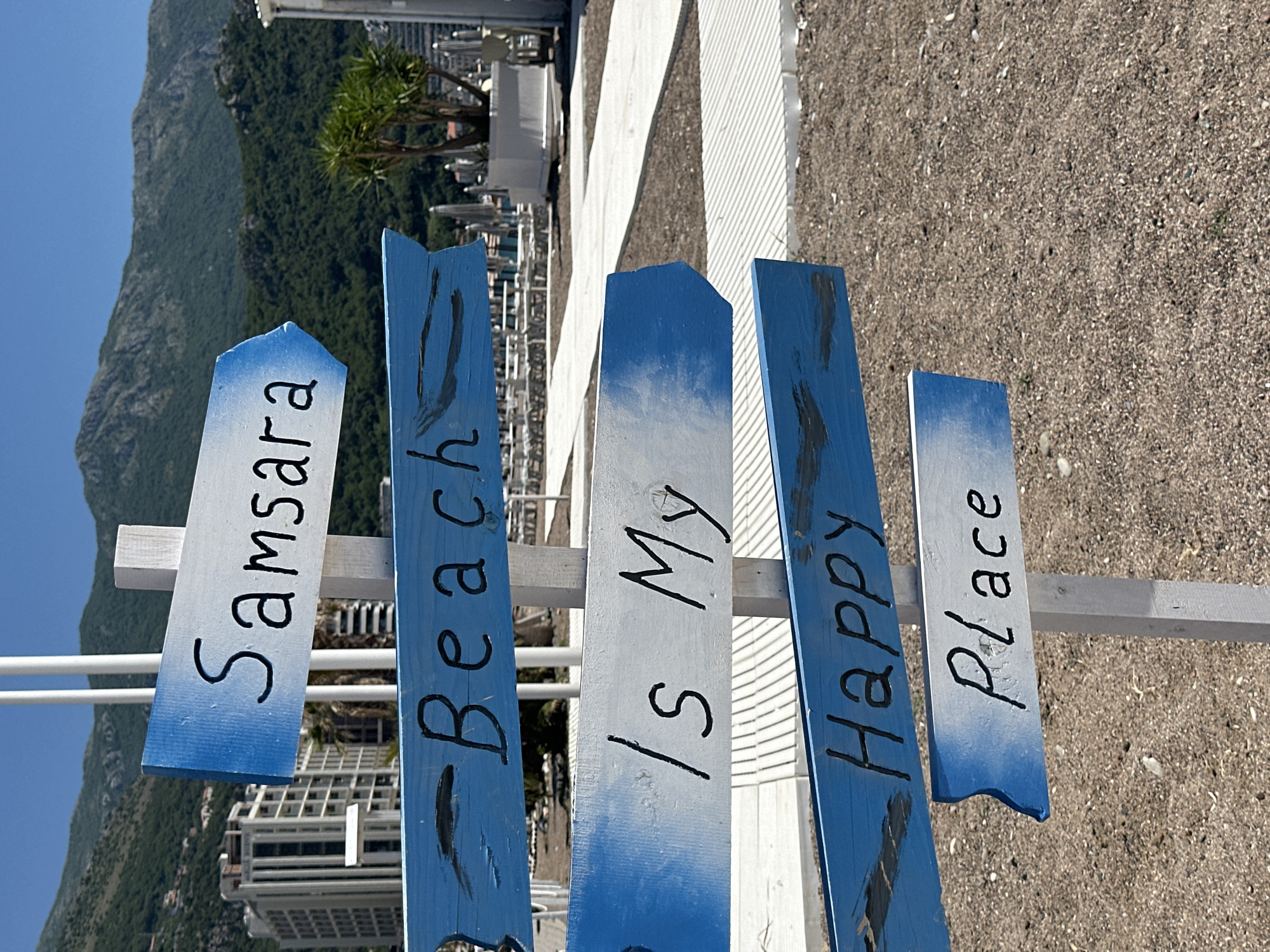 The image shows a sign at Samsara Beach in Boreti, Montenegro. The sign consists of five wooden planks painted in blue and white, with the words "Samsara Beach is my happy place" written on them in black. The sign is attached to a white wooden post, and the background includes a pebbled beach, a white boardwalk, buildings, a mountain, and a clear blue sky. The image is rotated to show the sign in an upright position.