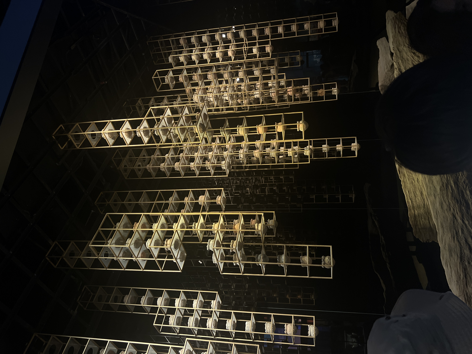 This image depicts an art installation in a dimly lit indoor space, likely a museum or gallery, in Yuzhong District, China. The primary subjects are numerous golden, rectangular frames suspended from the ceiling, creating vertical columns. Each frame contains multiple shelves, and on each shelf sits a small, light-colored, ceramic object, possibly vases or bowls, with varying textures and shapes. The objects are illuminated by spotlights, casting shadows and highlighting their intricate details. The background is predominantly black, with some areas reflecting the golden frames, suggesting the presence of mirrored surfaces. In the foreground, parts of the lower display, possibly large rocks or sculptures, are visible, along with the silhouettes of several people observing the installation. The overall impression is one of a curated exhibition designed to showcase these ceramic pieces in a dramatic and visually striking manner.