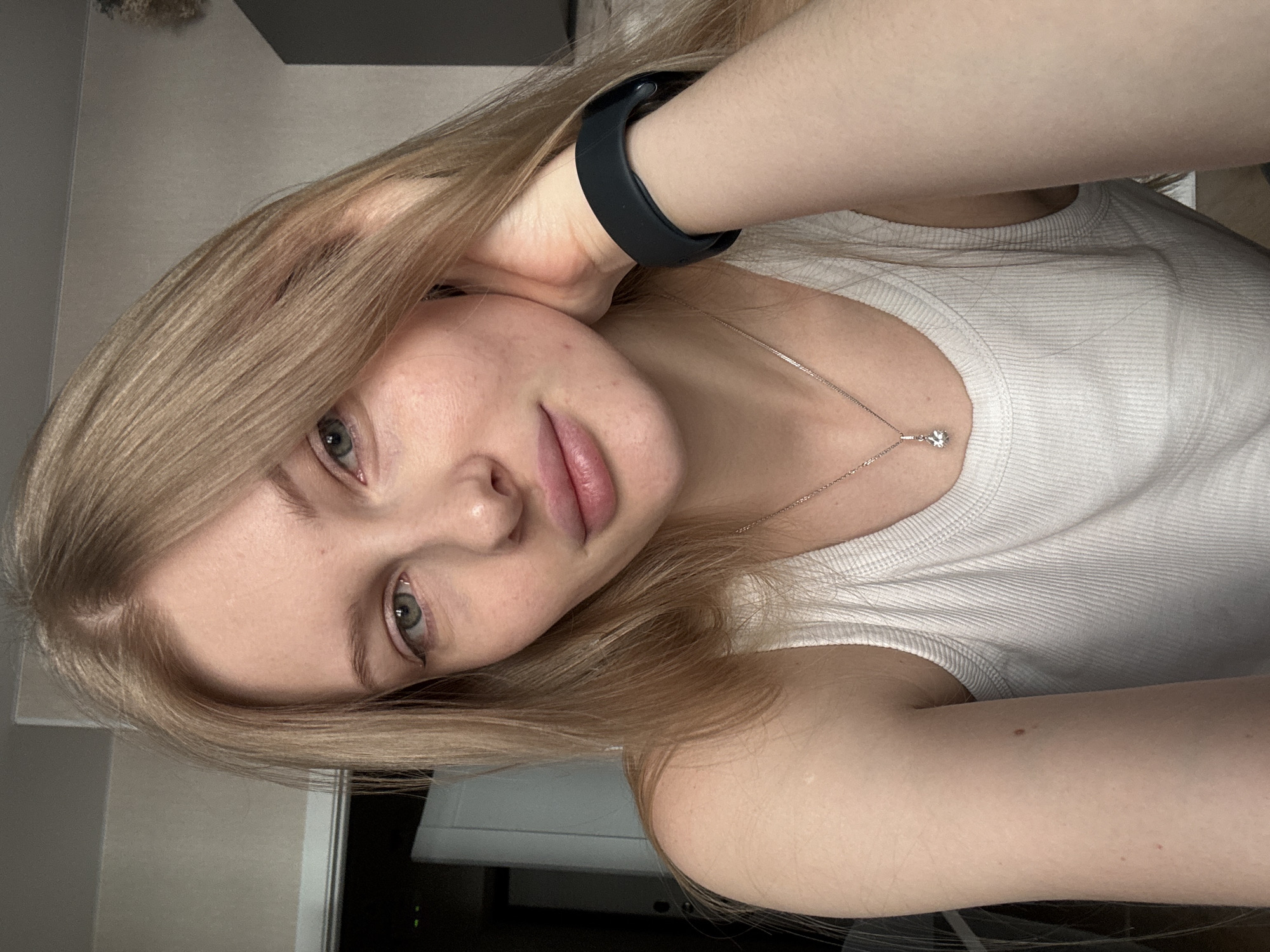A selfie, taken indoors, shows a fair-skinned young woman with light brown to blonde hair. She wears a white tank top and a black wristband. A silver necklace with a pendant is visible. The background is out of focus, appearing to show a wall and a portion of furniture. There is no evidence to suggest the location is Perm, Russia.