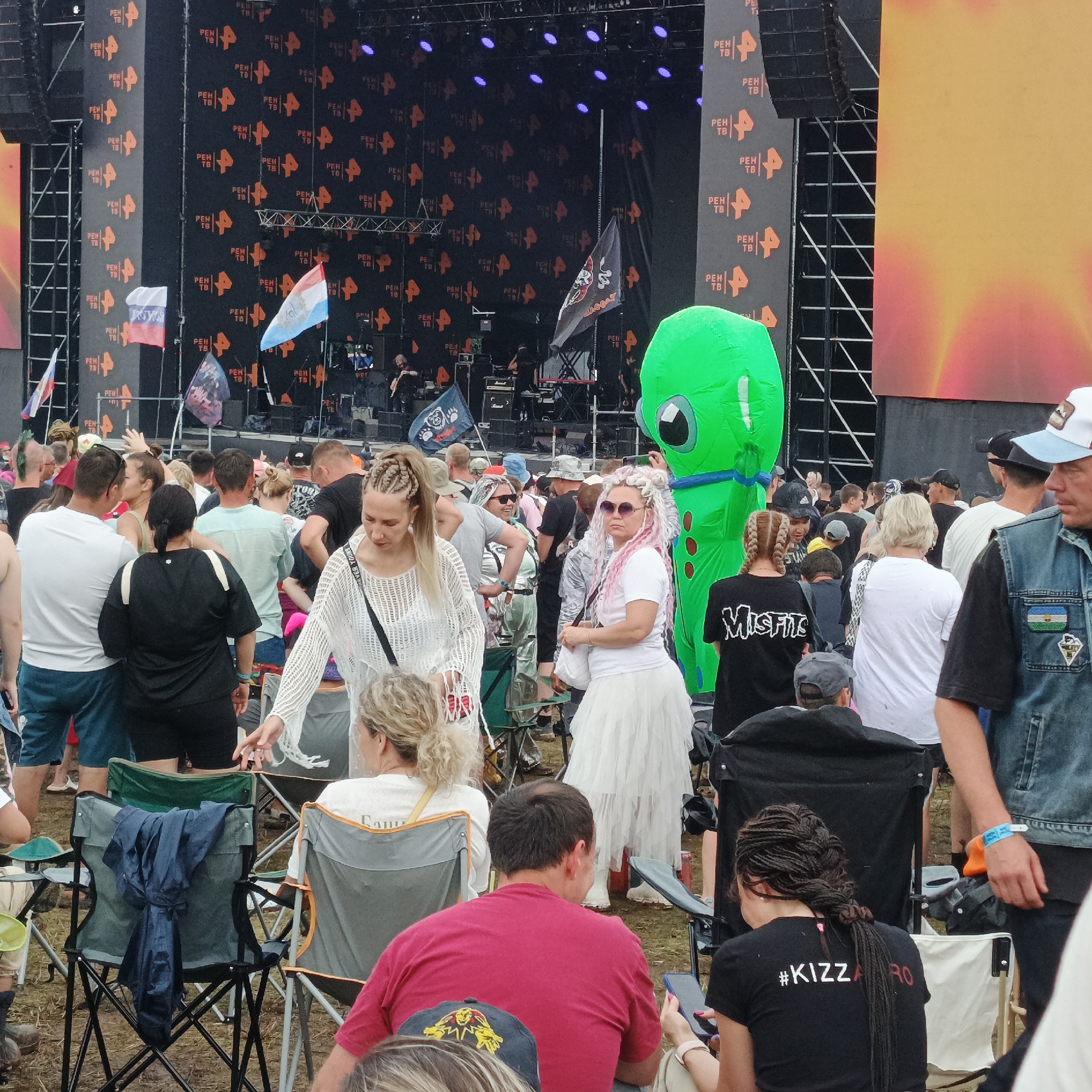 The image captures a scene at an outdoor music festival in Lagunovo, Russia. In the background, a large stage is set up with a screen displaying orange and black patterns featuring the logo "PEN TB". Several flags are visible, including the Russian flag and another with a blue and white design. A giant green inflatable alien-like figure stands to the right of the stage.
In the foreground, a crowd of people is gathered, many seated in camping chairs. The attendees appear to be enjoying the event. One woman in the foreground is wearing a white, crochet-like top and has blonde hair styled in braids. Another woman is wearing a white dress and has pink and white hair. A man in a red shirt is visible, as is a person in a black t-shirt with the text "#KIZZARO" on the back. The ground is covered with dirt and grass, indicating an outdoor venue. The overall atmosphere seems festive and relaxed, with people dressed casually for the event.