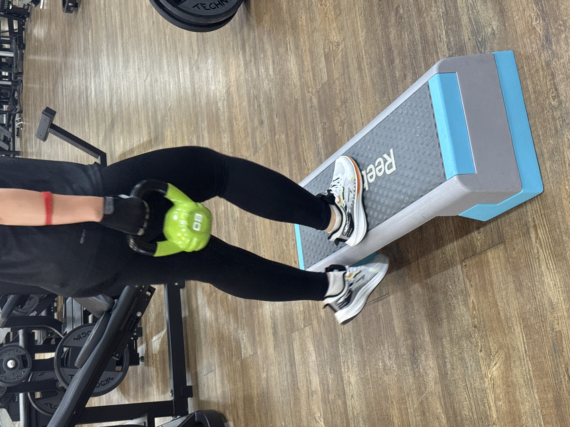 An individual is performing a step-up exercise on a grey and blue aerobic step. The person is wearing black leggings, white athletic shoes with black and orange accents, and black workout gloves. They are holding a bright green kettlebell with their right hand. The exercise step is placed on a wooden floor in what appears to be a gym. In the background, weight machines and weight plates are visible, suggesting a fitness facility. The individual's left foot is on the step, and their right foot is on the floor. The scene is lit by artificial lighting, typical of an indoor environment. There is no indication of the location being in the city of Chaikovsky, Russia, as the image lacks any specific geographical markers or identifying features.