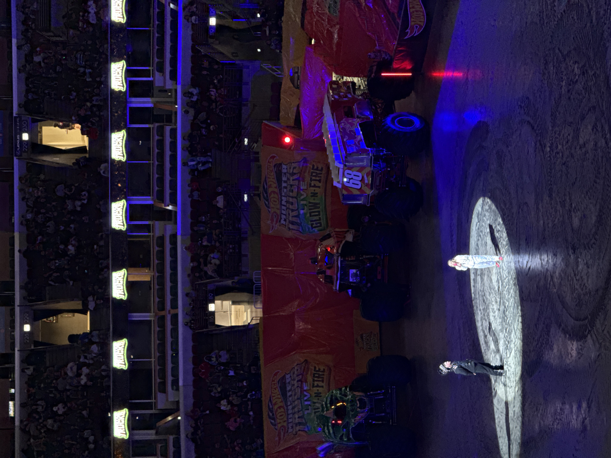In Chicago, Illinois, United States of America, a monster truck show is taking place in an indoor arena. The floor of the arena is covered in dirt and dust, with tire tracks visible. Two monster trucks are prominently displayed on the floor, one painted green with a skull design, and the other red and blue with the number "68" on its side. In the foreground, two individuals stand illuminated by spotlights. One, dressed in a black uniform with a patterned hat, stands to the left, while the other, in light-colored clothing, stands to the right. The arena is filled with spectators seated in tiered stands, bathed in the purple and blue lighting of the event. Banners with the "Hot Wheels Monster Trucks Live Glow-N-Fire" logo hang on the back wall. Neon signs that read "TRUCKS" are visible above the seating areas. The overall atmosphere appears to be one of excitement and entertainment.