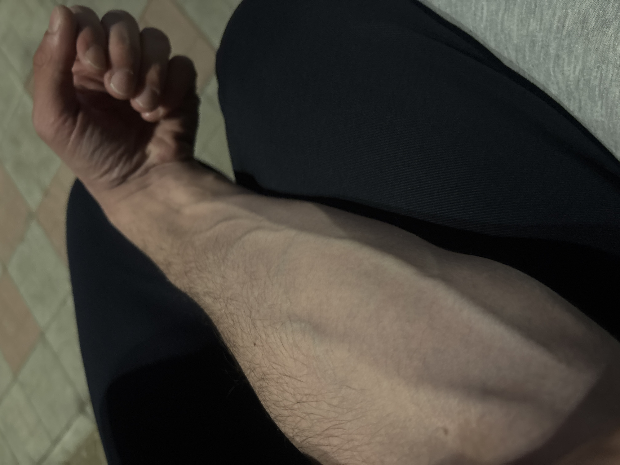 The image depicts a person's forearm and hand resting on what appears to be their lap. The forearm displays prominent veins. The hand is clenched into a fist. The person is wearing dark clothing, with a lighter-colored garment visible in the background. The setting appears to be indoors, near a tiled floor. There is no information to confirm a location in Yerevan, Armenia.