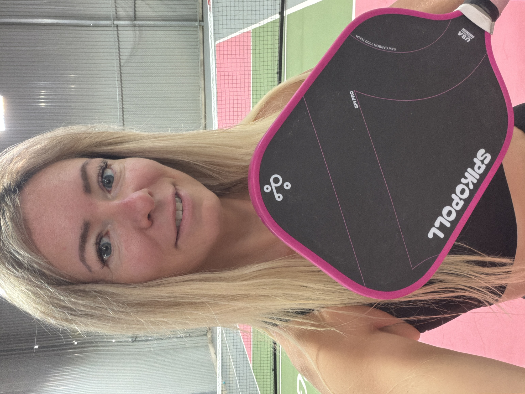 An individual with blonde hair is holding a pink and black pickleball paddle in what appears to be an indoor pickleball court. The person is smiling and looking towards the camera. The court has green and pink playing surfaces and a net in the background. The walls of the indoor facility are made of corrugated metal. There is no indication of the city of Da Lat, Vietnam, in the image.