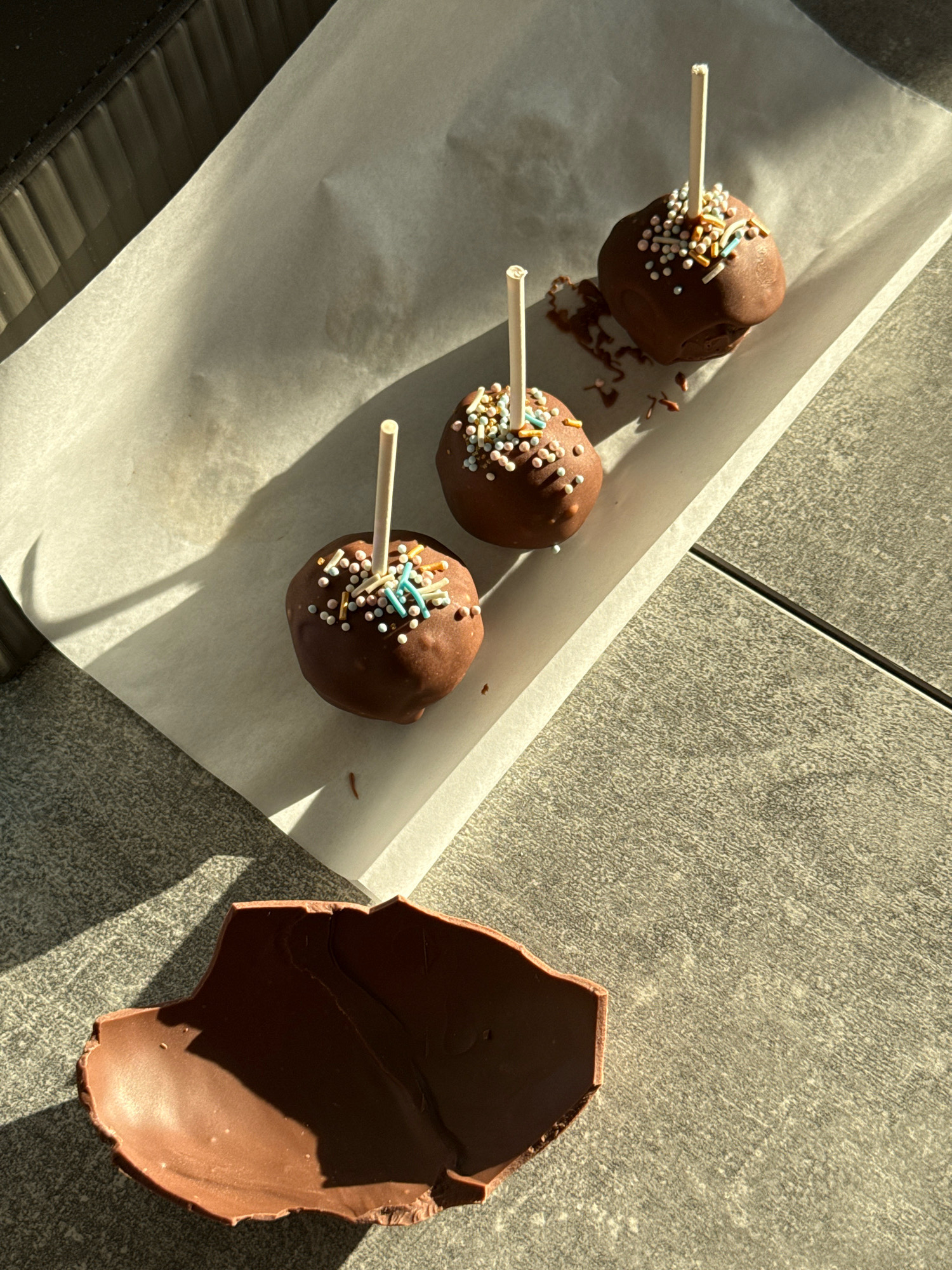 Three chocolate cake pops with colorful sprinkles are arranged on white parchment paper. A broken piece of a chocolate Easter egg lies in the foreground, casting a shadow. The scene is set outdoors on a textured, gray surface, possibly a patio or table, bathed in sunlight. The overall composition suggests a sweet, possibly festive, occasion. There are no people or animals present, and no specific landmarks in Lyubertsy, Russia, are identifiable.