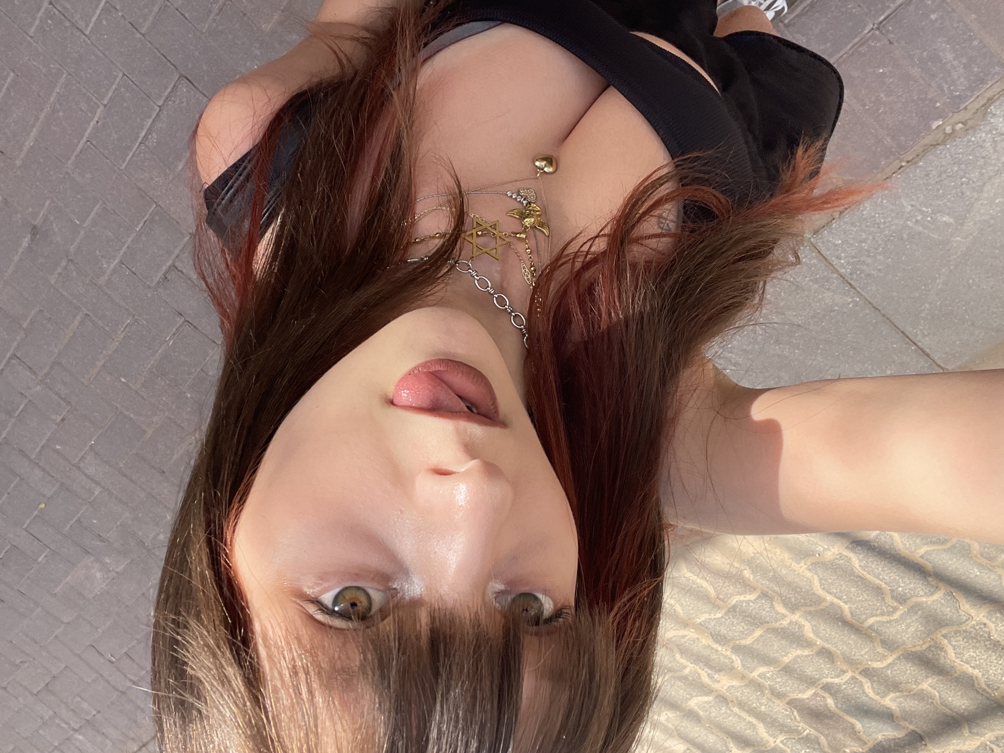 A selfie taken from a low angle depicts a young woman with light skin, hazel eyes, and brown hair with red highlights. Her hair is styled with bangs covering part of her forehead, and her tongue is slightly visible. She is wearing a black top that exposes her cleavage, and multiple necklaces with charms are visible. The background appears to be a paved area, possibly a sidewalk, with a repeating geometric pattern. The presence of sunlight suggests the photo was taken outdoors during the day. The location in the city of Raanana, Israel, cannot be confirmed from the visual data alone.