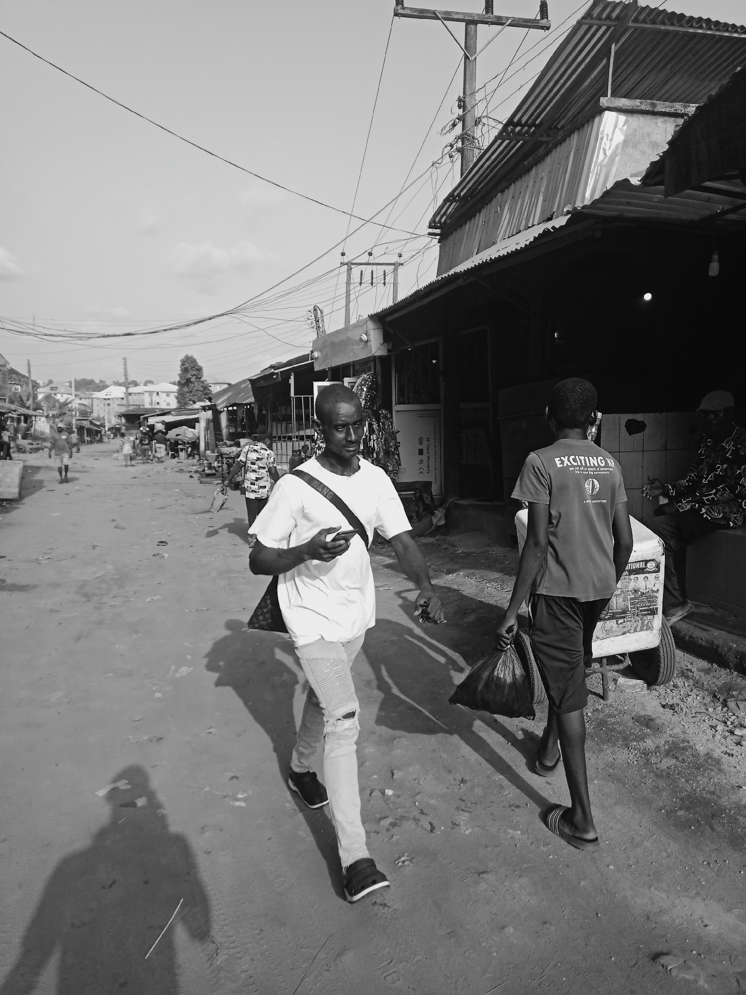 A black and white photograph shows a street scene in Onitsha, Nigeria. A young man in a light-colored t-shirt and distressed jeans walks towards the camera, holding a cell phone. He has a shoulder bag. To his right, another young man walks away from the camera, carrying a black plastic bag. He wears a t-shirt and shorts, and sandals. He is pulling a small white cart, possibly a mobile vendor unit. Other people are visible in the background, moving along the street. Buildings line the street, with corrugated metal roofing and shopfronts. Power lines crisscross above. The ground is unpaved and dusty.