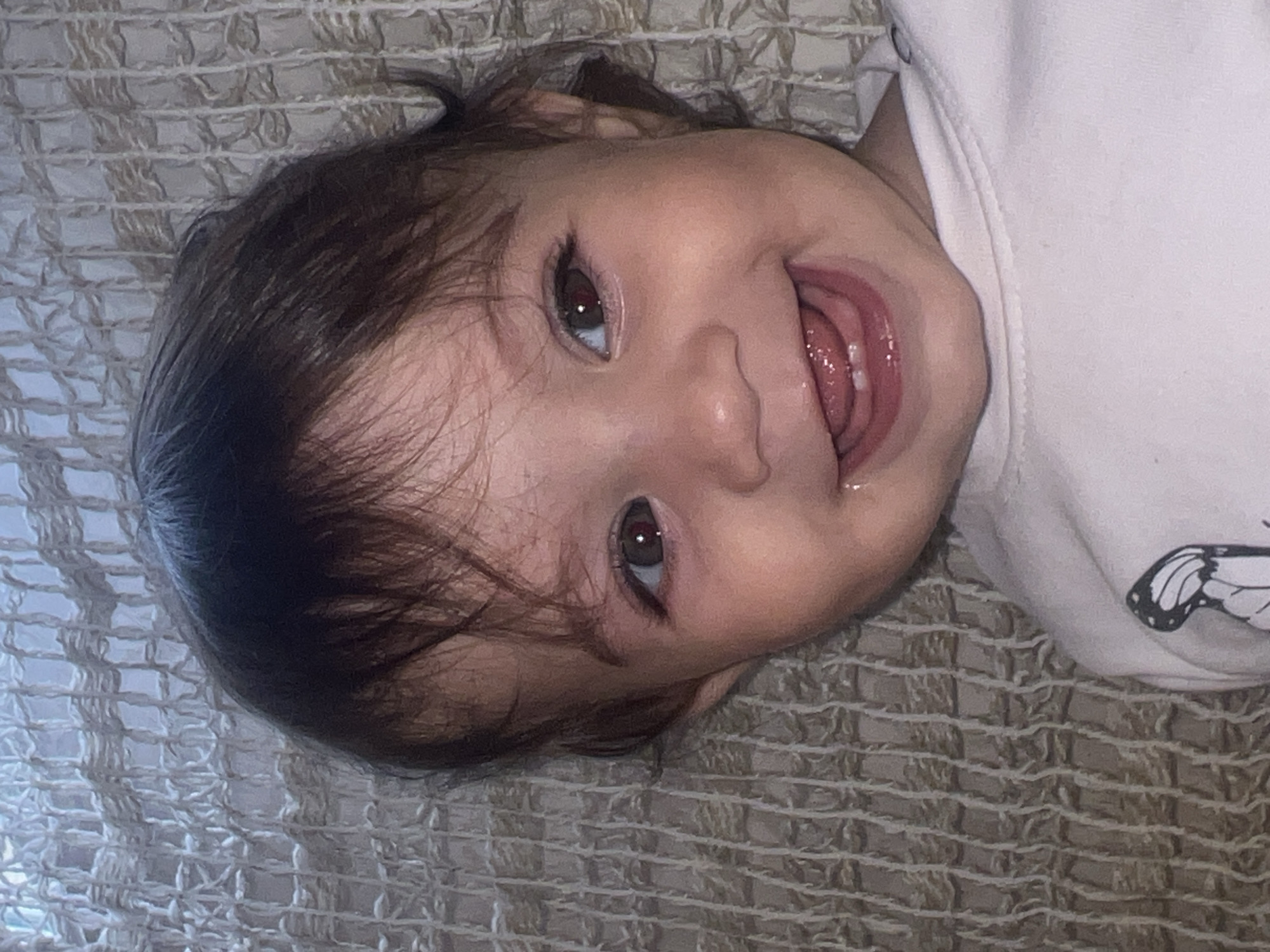 A close-up, eye-level shot depicts a baby with dark brown hair and light skin, lying on a textured, beige-colored surface. The baby is smiling, with its mouth open, revealing a few teeth and its tongue. The baby is wearing a white shirt with a butterfly design visible on the right side of the frame. The lighting is diffused, casting soft shadows across the scene. The background is the same textured surface, providing a neutral setting. The location in Elena, Bulgaria, is not discernible from the image.