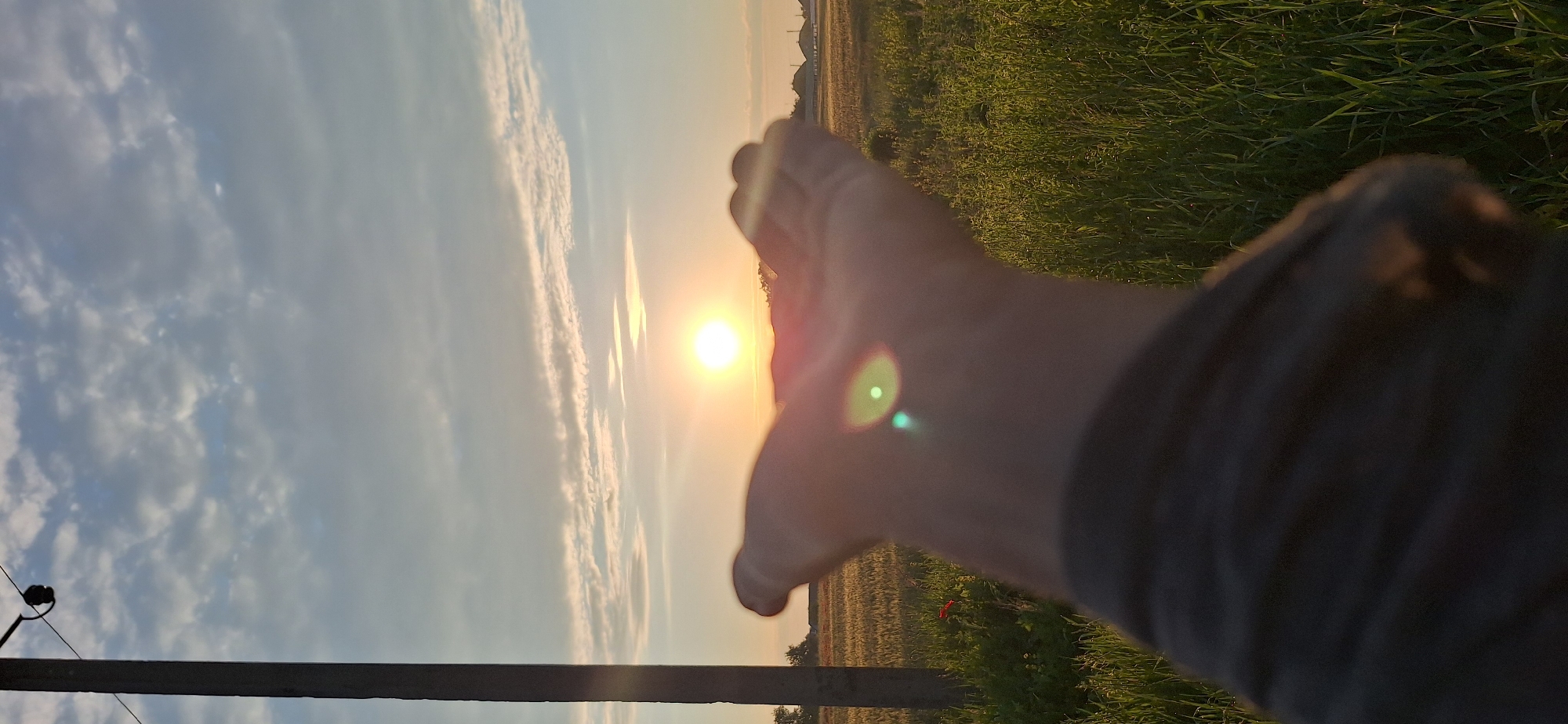 An outstretched hand is captured in the foreground of this vertical, eye-level, medium shot taken outdoors. The hand is angled upwards, cupping the air, with a bright sun positioned directly above it. The sun is emitting a lens flare that creates a green circular reflection with a smaller green dot in its center. Behind the hand, a field of tall green grass extends to the horizon. In the far distance, several buildings with domed roofs are visible. To the left of the frame, a utility pole stands tall, and further to the left, a field of golden crops can be seen, with a single red poppy flower in bloom. The sky above is a soft blue, filled with wispy white clouds. The lighting suggests it is either sunrise or sunset.