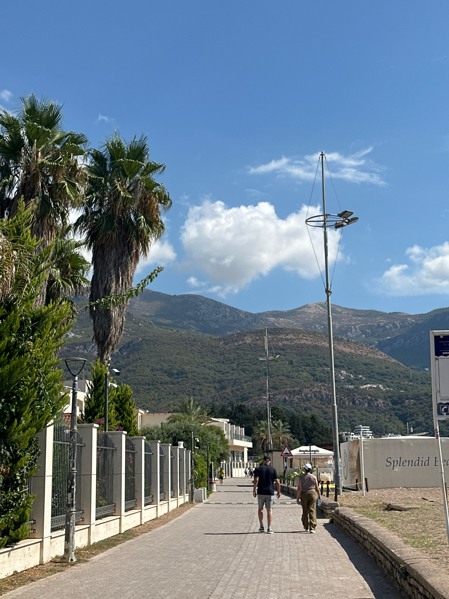 This image captures a scenic view of the promenade in Boreti, Montenegro, on a clear day. In the foreground, a paved walkway stretches into the distance, lined with a decorative fence and lush green foliage, including prominent palm trees. Two individuals, a man and a woman, are walking away from the camera on the walkway. The man wears a dark t-shirt and grey shorts, while the woman is dressed in a light-colored t-shirt and olive-green pants.
Further down the promenade, more people can be seen walking, and buildings with white facades and balconies are visible. Beyond the buildings and vegetation, a large, imposing mountain range covered in dense green forest dominates the background. The sky is a vibrant blue, punctuated by a few white, fluffy clouds. A tall lamp post with multiple lights stands near the center of the image, and a sign with the text "Splendid Bea" is visible on the right side of the promenade, suggesting the proximity of a beach. The overall scene depicts a relaxed atmosphere in a coastal town with a mountainous backdrop.