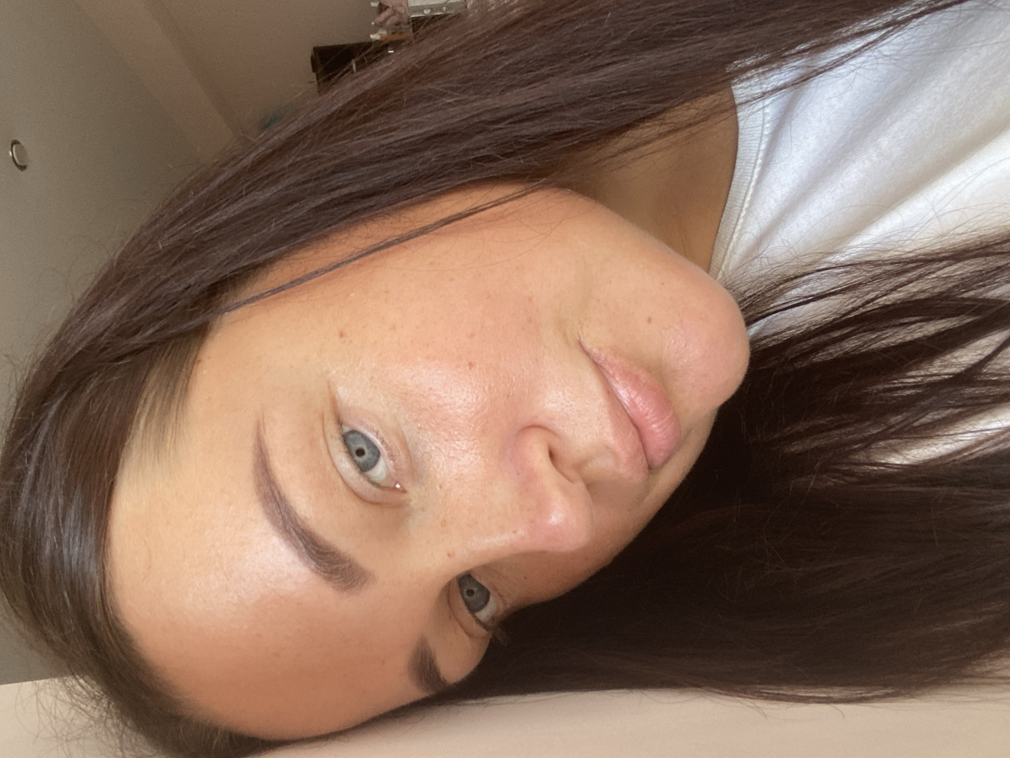 The image is a close-up, outdoor, selfie-style portrait of a woman with long, dark brown hair and blue eyes. She is leaning her head against a light-colored wall, and her face is angled towards the camera. She appears to be wearing a white t-shirt. The background is softly blurred, with hints of indoor lighting and possibly furniture. There are no discernible landmarks or context to suggest the location in Sochi, Russia.