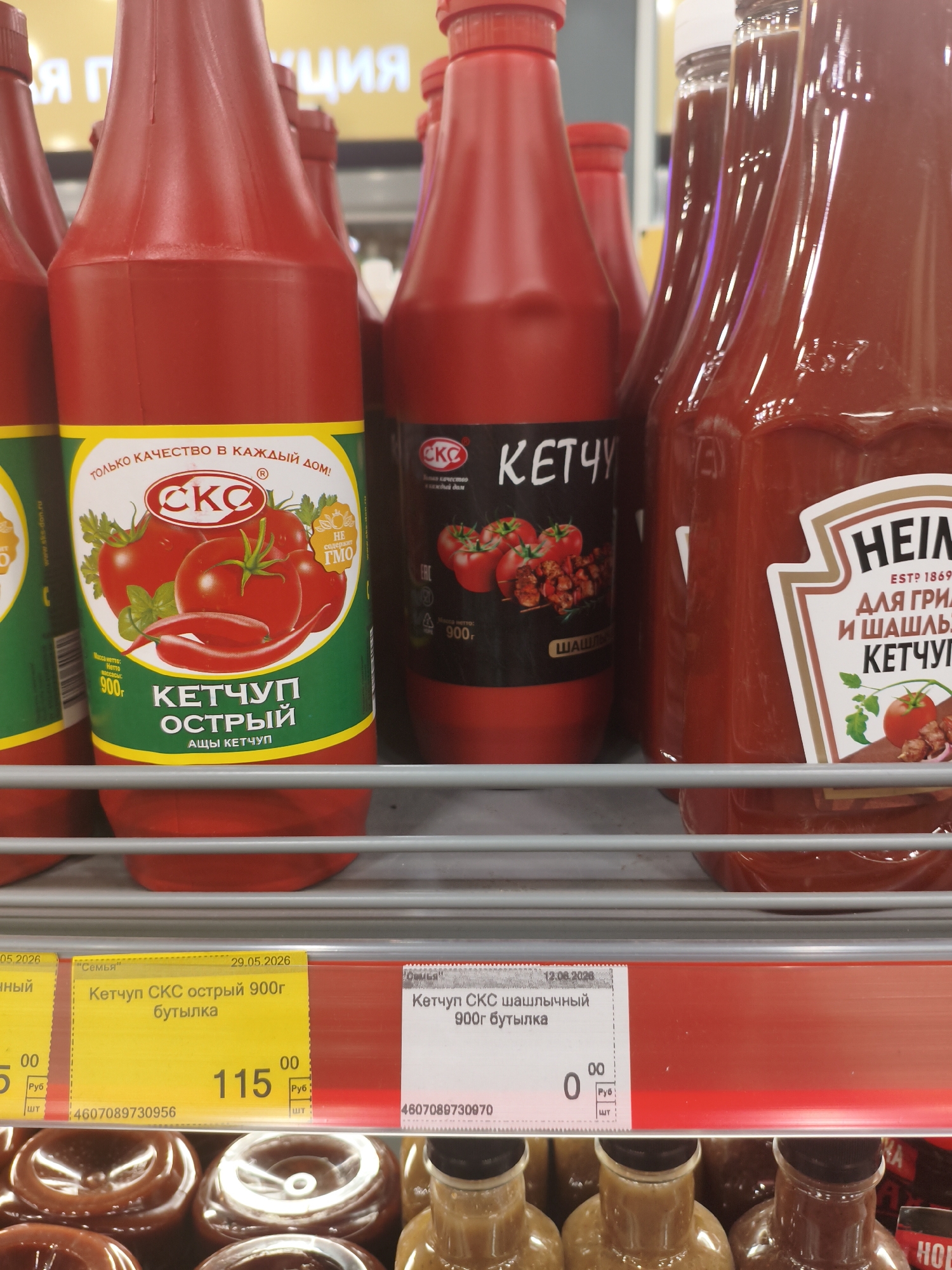 This image shows a shelf in a supermarket displaying several bottles of ketchup. In the foreground, there are multiple bottles of "CKC" brand ketchup, with one clearly labeled "КЕТЧУП ОСТРЫЙ" (Spicy Ketchup). To the right, there is a bottle of "HEINZ" ketchup, described as "ДЛЯ ГРИЛЬ И ШАШЛЫК" (For Grill and Shashlik). Below the ketchup bottles, there are price tags visible. One price tag shows "115" rubles for "Кетчуп СКС острый 900г бутылка" (CKC Spicy Ketchup 900g bottle), with an expiration date of 29.05.2026. Another price tag, to the right, is for "Кетчуп СКС шашлычный 900г бутылка" (CKC Shashlik Ketchup 900g bottle) and shows a price of "0.00" rubles with an expiration date of 12.08.2026. The environment appears to be a retail setting with fluorescent lighting. The image is a close-up, eye-level shot of the products on the shelf.