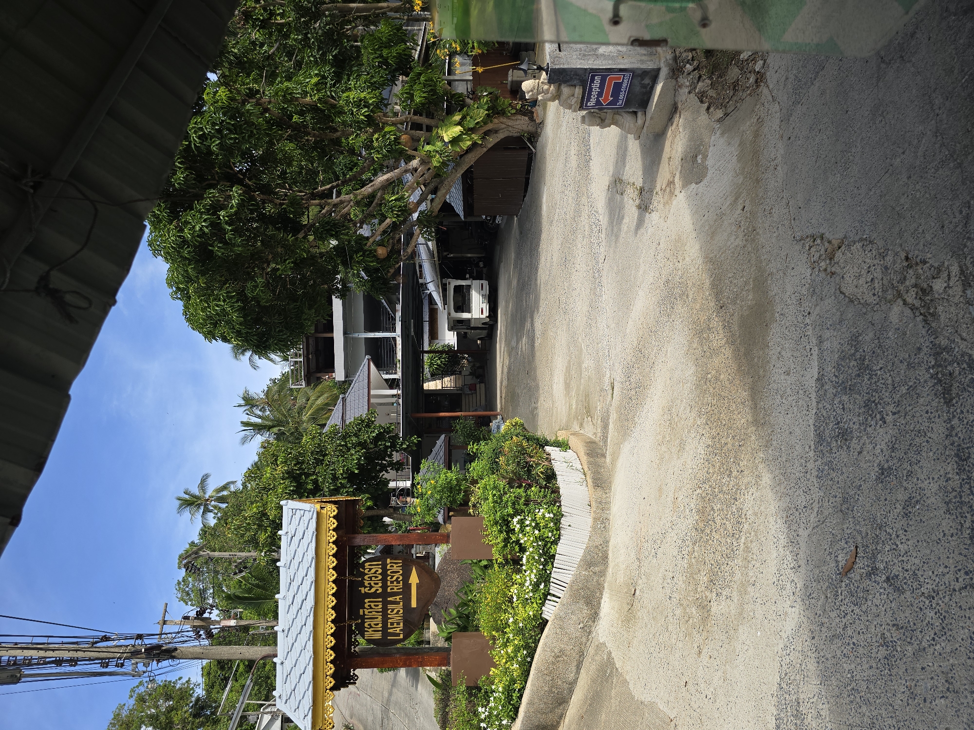 A resort entrance is depicted in the image, featuring a wooden sign with Thai script and "LAEMSILA RESORT" written below it, along with a yellow arrow pointing to the right. The sign is surrounded by a garden of green plants and white flowers. To the right of the sign, a paved driveway curves upwards. In the background, a white van is parked in front of a building complex. Further to the right, a sign indicates "Reception" with a phone number and a red arrow. The environment is lush with tropical vegetation, including palm trees, and the sky is blue with some clouds. The overall impression is that of a tropical resort entrance under a bright sky.