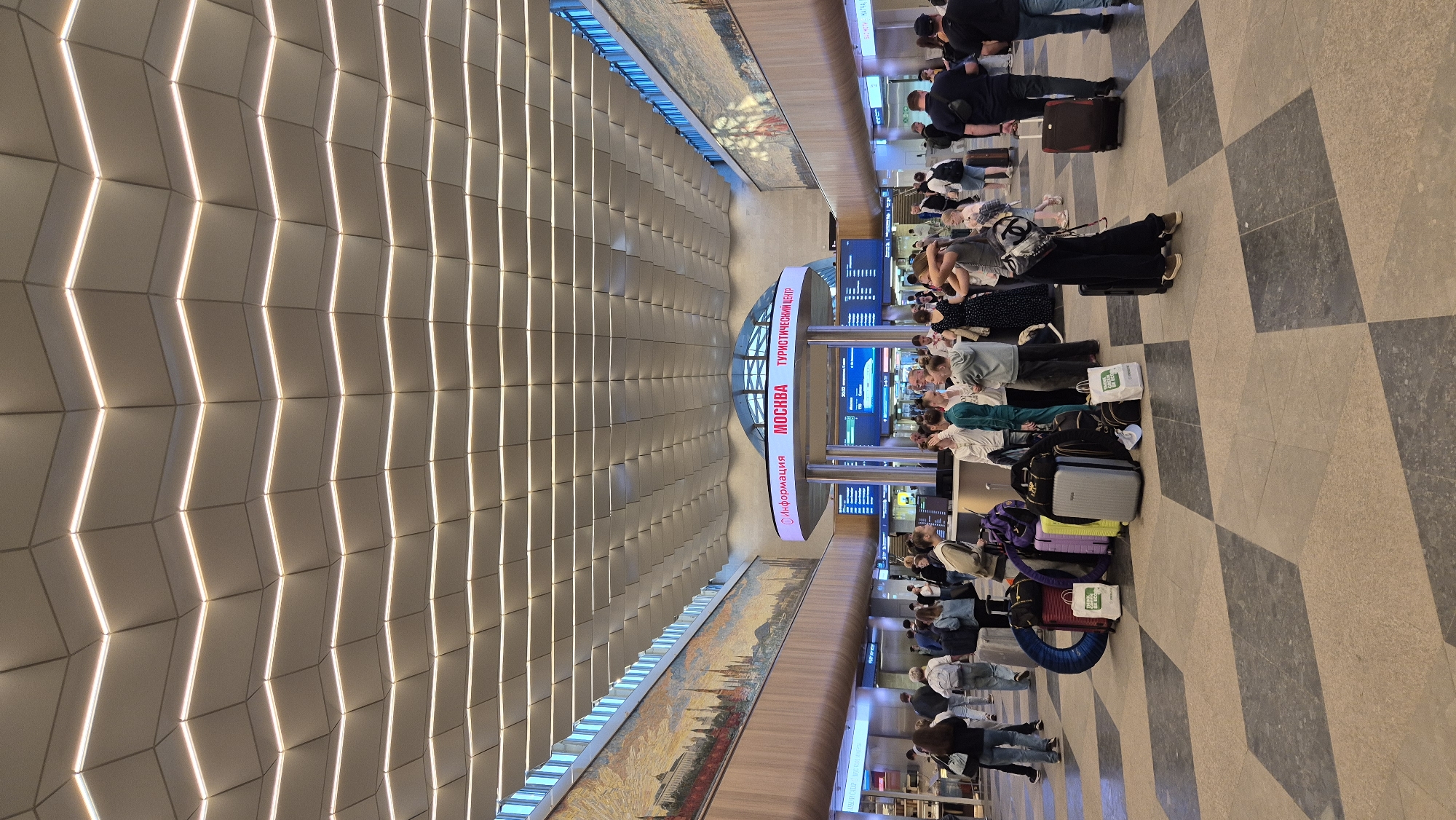 The image depicts the interior of a modern, spacious transportation hub, likely a train station or airport terminal, in Moscow, Russia. The architecture features a distinctive, undulating ceiling with integrated linear lighting that creates a dynamic visual effect. Large-scale murals depicting cityscapes adorn the upper walls on either side of the hall. In the central area, a circular information kiosk displays "МОСКВА" (Moscow) and "ТУРИСТИЧЕСКИЙ ЦЕНТР" (Tourist Center), indicating its function. Digital screens showing departure or arrival information are visible behind the kiosk.
A diverse group of people, including adults and children, are present in the space, many with luggage. Several individuals are standing in clusters, conversing, while others are walking or waiting. A prominent collection of suitcases, including hard-shell cases, duffel bags, and a distinctive blue inflatable ring, is arranged on the checkered floor in the foreground. The floor itself is a patterned design of dark grey and beige diamond shapes. The overall atmosphere suggests a busy, transitional space associated with travel.