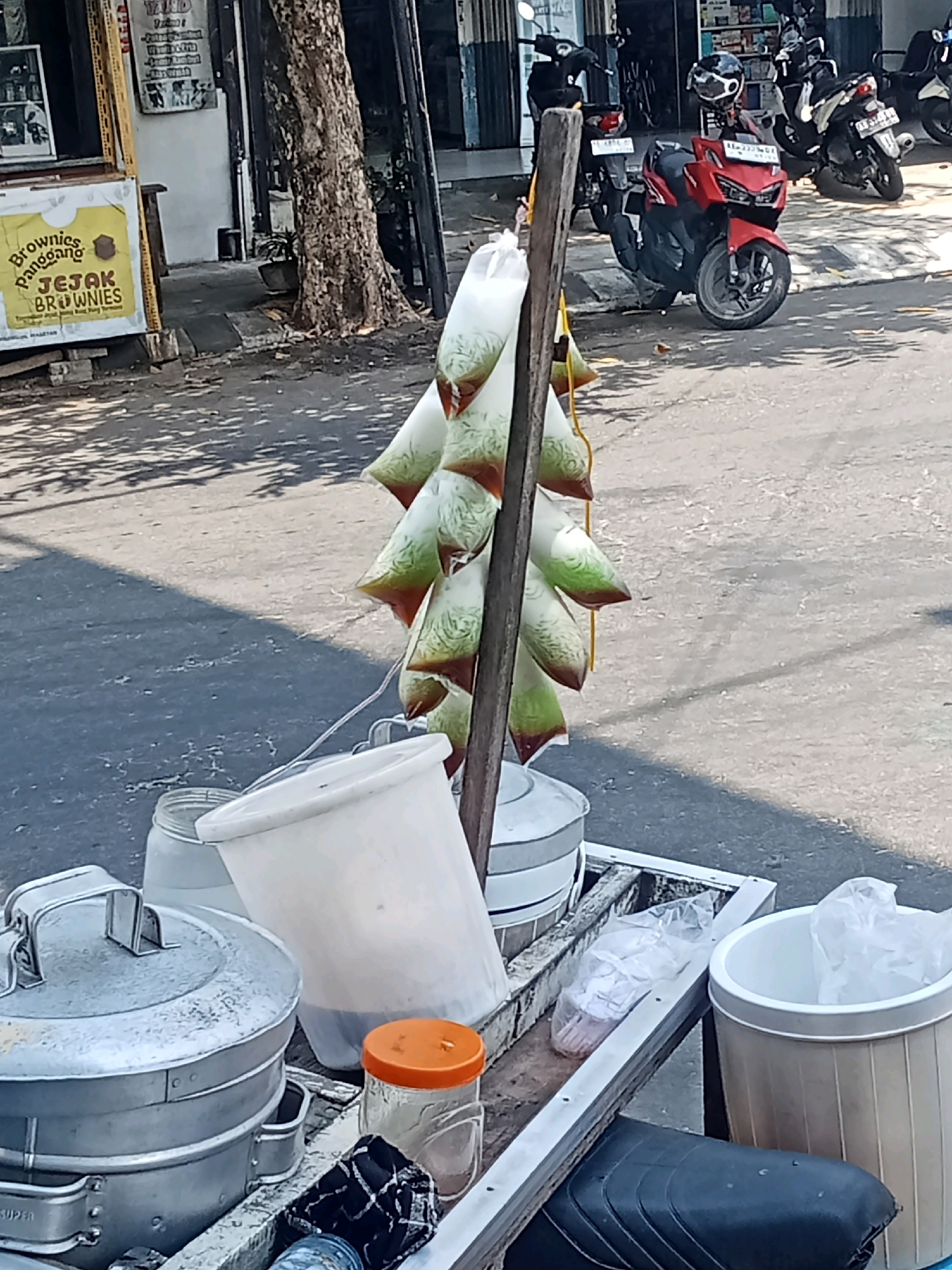 The image shows a street scene in Sukowinangun, Indonesia, with a focus on a food cart. The cart is laden with various items, including large metal containers, a white bucket, and several plastic bags filled with a green and brown liquid, hanging from a wooden pole. An orange-lidded jar is also visible on the cart. In the background, several motorcycles are parked, and a tree with a rough bark stands prominently. A storefront with signage is visible behind the tree, and the ground is paved asphalt, with shadows from trees cast across it. The overall impression is of a bustling urban environment where street food is being sold.