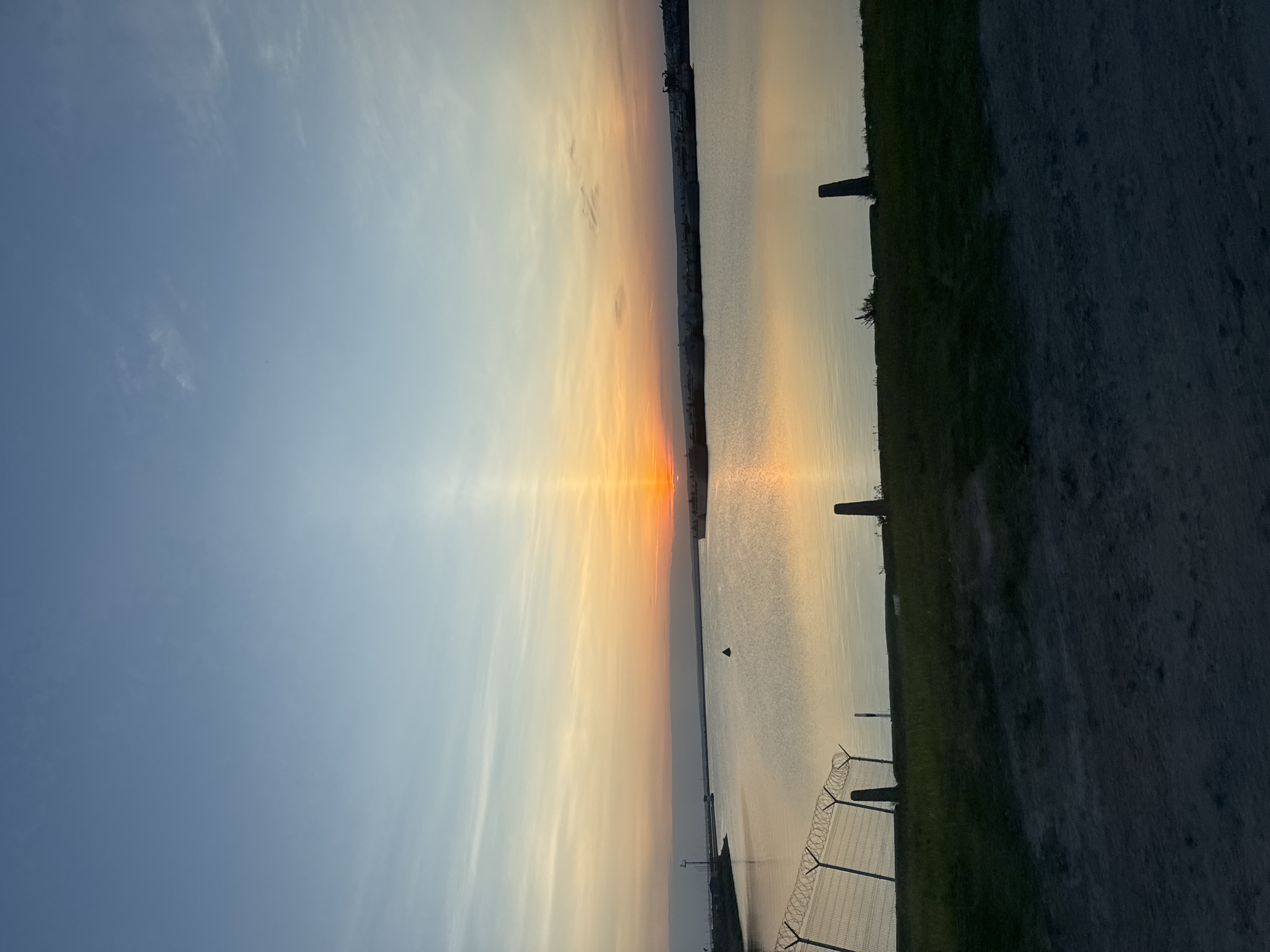 The image shows a landscape at sunset, likely near a body of water. The sky transitions from blue to orange and yellow hues near the horizon. A thin line of land with buildings is visible on the horizon. The water reflects the colors of the sky. A fence with barbed wire runs along the right side of the image, adjacent to a patch of green grass. A paved area is also on the right side. Considering the query includes Chaikovsky, Russia, the landscape might be near the Kama River, the Votkinsk Reservoir, or a similar body of water in or near the city. There are no people visible in the image. The image focuses on the natural beauty of the sunset over water.